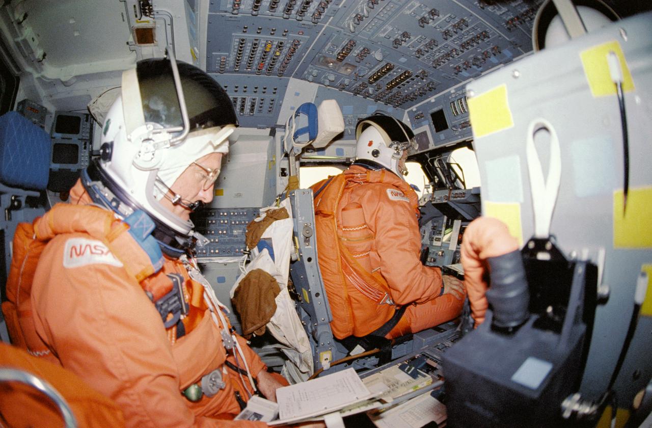 STS029-24-004 (18 March 1989) --- STS-29 crewmembers, wearing launch and entry suits (LESs) and launch and entry helmets (LEHs), review checklists on Discovery, Orbiter Vehicle (OV) 103, flight deck. Commander Michael L. Coats is seated at the forward flight deck commanders station with Mission Specialist (MS) James F. Buchli on aft flight deck strapped in mission specialist seat. OV-103 makes its return after five days in space. Note color in forward windows W1, W2, W3 caused by friction of entry through the Earth's atmosphere. Personal Egress Air Pack (PEAP) is visible on pilots seat back.