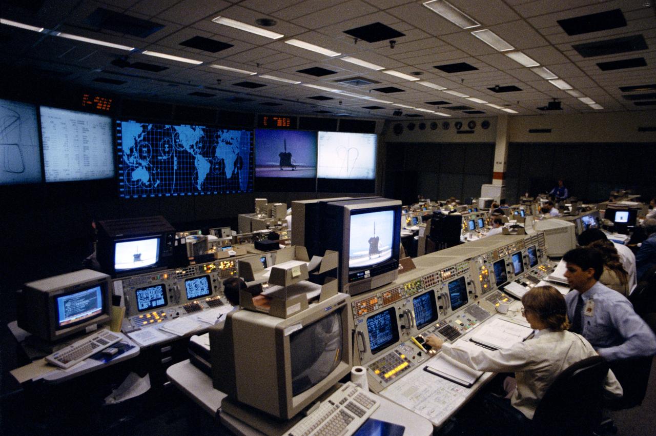 As STS-30 ends its mission with the landing of Atlantis, Orbiter Vehicle (OV) 104, at Edwards Air Force Base (EAFB), California, flight controllers monitor screens at their consoles in JSC Mission Control Center (MCC) Bldg 30. In the foreground is the Maintenance, Mechanical Arm, and Crew Systems Engineer (MMACS) console with R. Kevin McCluney studying data readouts. On the front visual displays are the tracking map and a tail view of OV-104 as it comes to a stop on EAFB concrete runway 22.