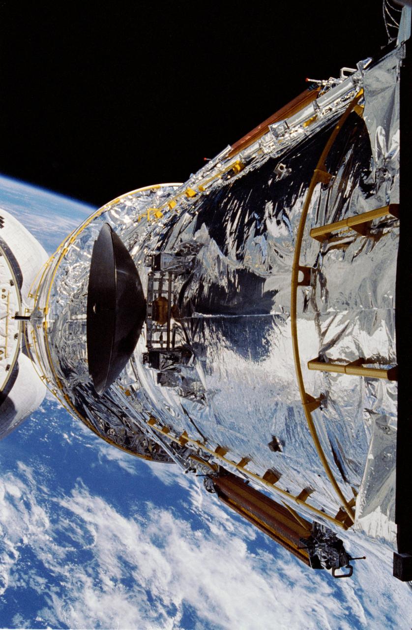 The Hubble Space Telescope (HST) is raised above the payload bay (PLB) in low hover position during STS-31 checkout and pre-deployment procedures aboard Discovery, Orbiter Vehicle (OV) 103. Stowed along the HST Support System Module (SSM) are the high gain antenna (HGA) (center) and the two solar arrays (one either side). In the background are the orbital maneuvering system (OMS) pods and the Earth's surface.