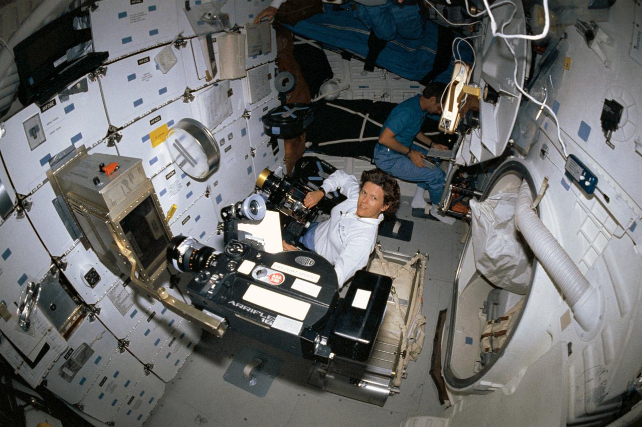 STS031-05-002 (24-29 April 1990) ---  A 35mm camera with a "fish eye" lens captured this high angle image on Discovery's middeck.   Astronaut Kathryn D. Sullivan works with the IMAX camera in foreground, while Astronaut Steven A. Hawley consults a checklist in corner.  An Arriflex motion picture camera records student ion arc experiment in apparatus mounted on stowage locker.  The experiment was the project of Gregory S. Peterson, currently a student at Utah State University.