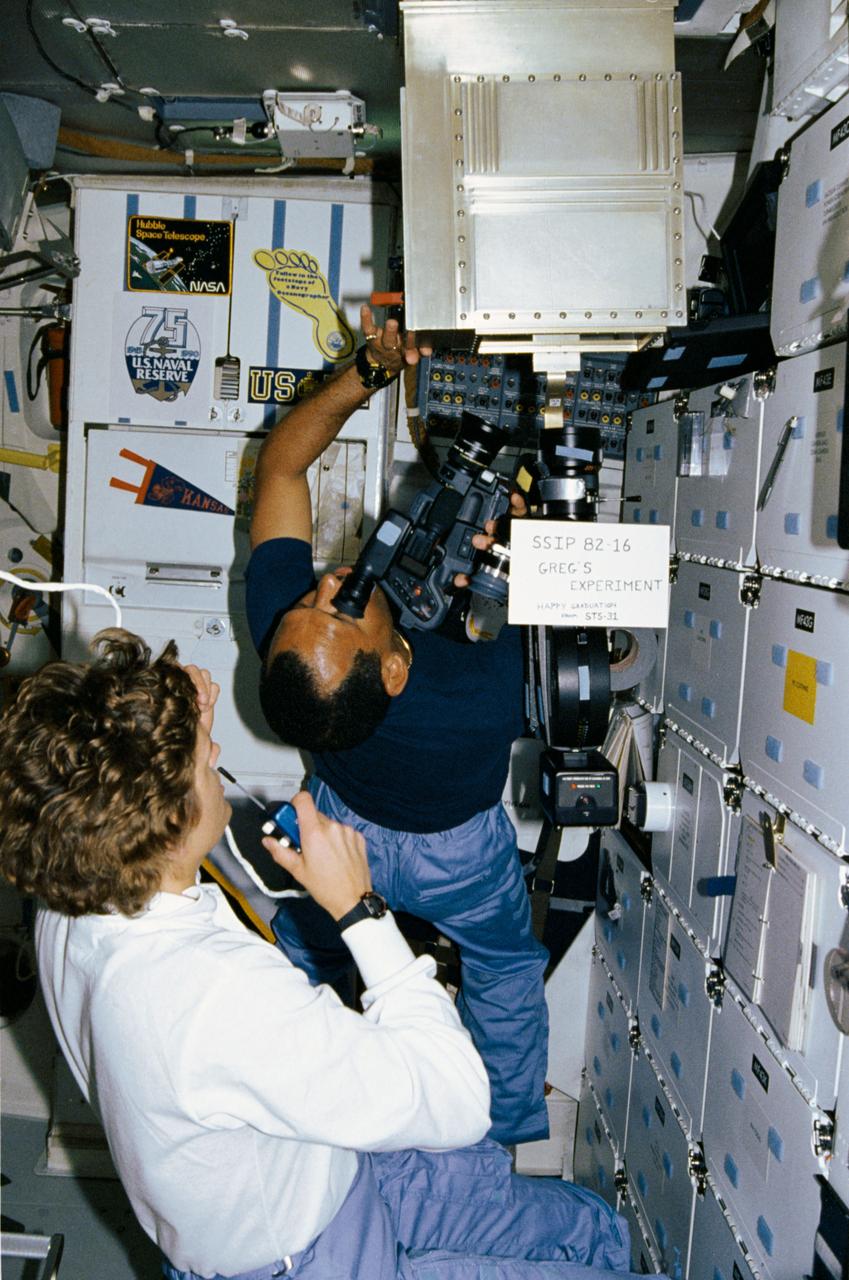 STS-31 Mission Specialist (MS) Kathryn D. Sullivan monitors and advises ground controllers of the activity inside the Student Experiment (SE) 82-16, Ion arc - studies of the effects of microgravity and a magnetic field on an electric arc, mounted in front of the middeck lockers aboard Discovery, Orbiter Vehicle (OV) 103. Pilot Charles F. Bolden uses a video camera and an ARRIFLEX motion picture camera to record the activity inside the special chamber. A sign in front of the experiment reads "SSIP 82-16 Greg's Experiment Happy Graduation from STS-31." SSIP stands for Shuttle Student Involvement Program. Gregory S. Peterson who developed the experiment (Greg's Experiment) is a student at Utah State University and monitored the experiment's operation from JSC's Mission Control Center (MCC) during the flight. Decals displayed in the background on the orbiter galley represent the Hubble Space Telescope (HST), the United States (U.S.) Naval Reserve, Navy Oceanographers, U.S. Navy, and University of Kansas.