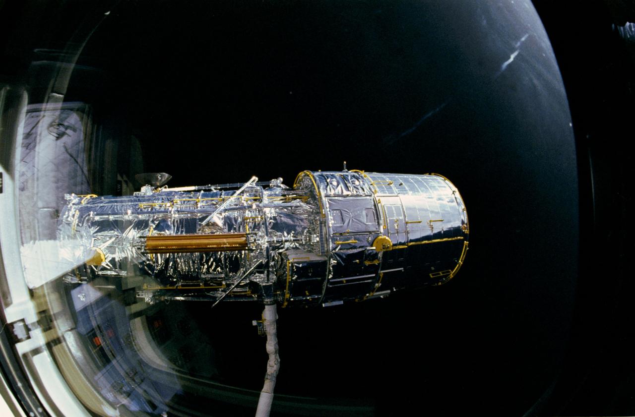 During STS-31 checkout, the Hubble Space Telescope (HST) is held in a pre-deployment position by Discovery's, Orbiter Vehicle (OV) 103's, remote manipulator system (RMS). The view, taken from the crew cabin overhead window W7, shows the starboard solar array (SA) panel (center) and two high gain antennae (HGA) (on either side) stowed along side the Support System Module (SSM) forward shell. The sun highlights HST against the blackness of space.