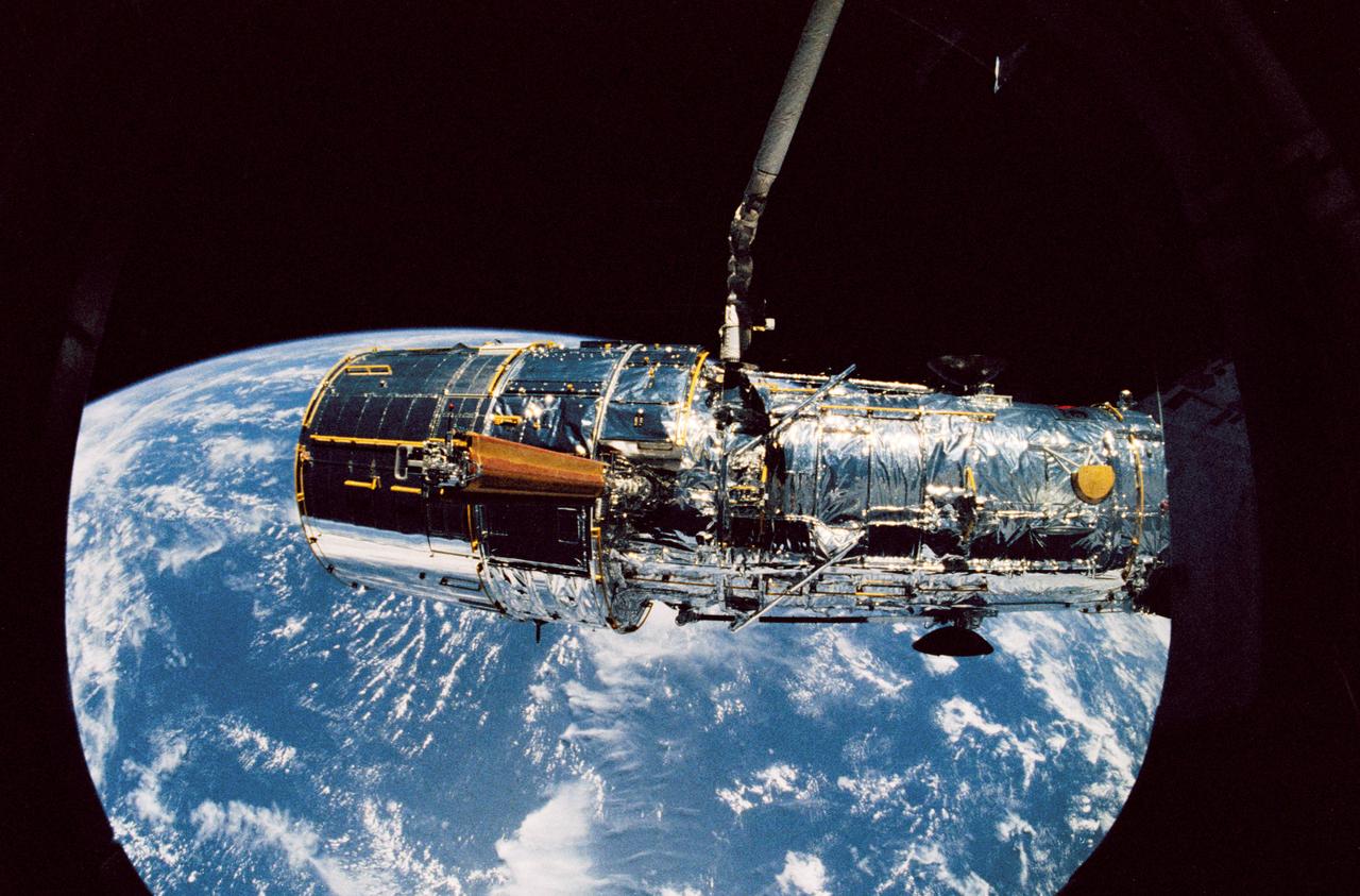 Held in appendage deploy position by Discovery's, Orbiter Vehicle (OV) 103's, remote manipulator system (RMS), the Hubble Space Telescope's (HST's) starboard solar array (SA) bistem cassette is released from its stowed position on the Support System Module (SSM) forward shell. The spreader bar & bistem begin to unfurl the SA wing. View was taken by an STS-31 crewmember through an overhead window & is backdropped against the surface of the Earth.