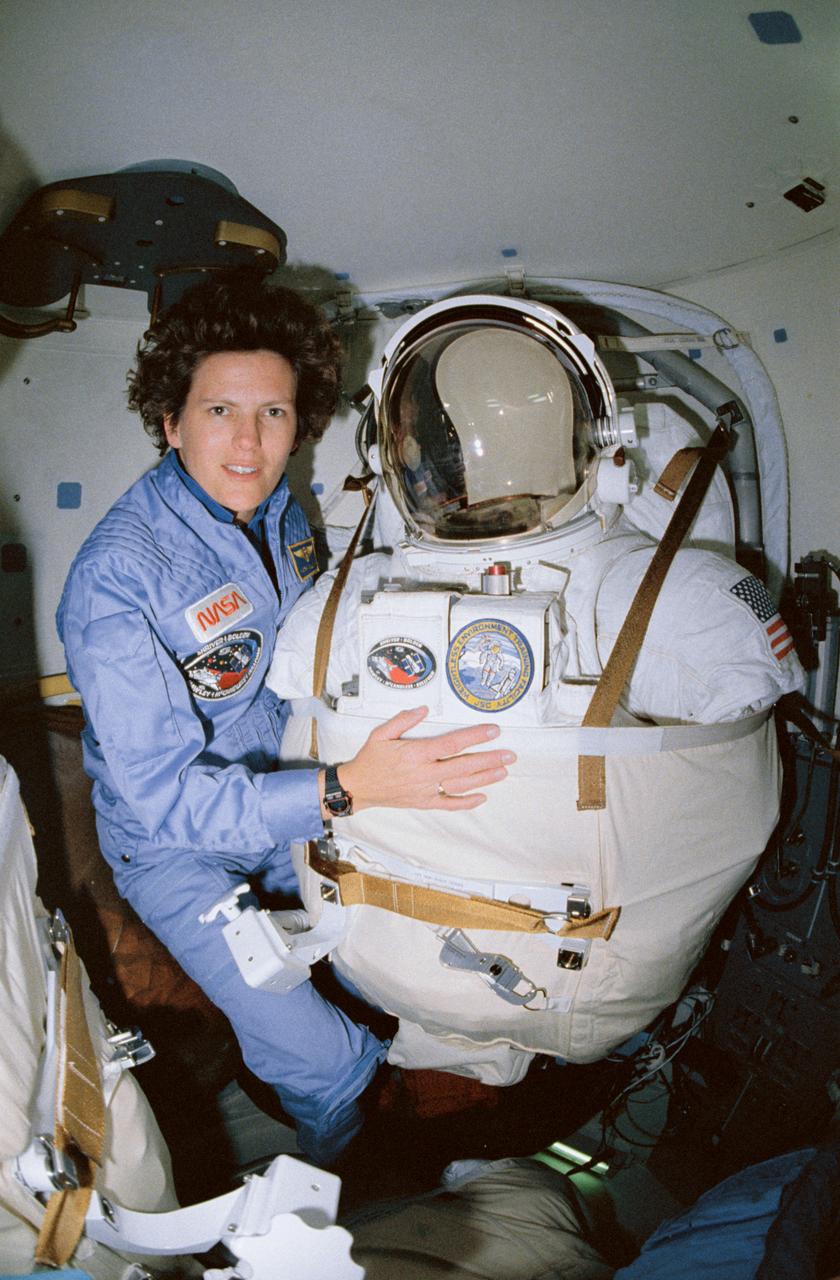 STS-31 Mission Specialist (MS) Kathryn D. Sullivan poses for a picture before beginning extravehicular mobility unit (EMU) donning procedures in the airlock of Discovery, Orbiter Vehicle (OV) 103. Sullivan will remove the lower torso restraint and don EMU which is supported on an airlock adapter plate (AAP). When suited, Sullivan will be ready for contingency extravehicular activity (EVA) in the event that problems arise with the Hubble Space Telescope (HST) deployment. Displayed on the front of the EMU are the STS-31 mission insignia and the JSC Weightless Environment Training Facility (WETF) insignia.