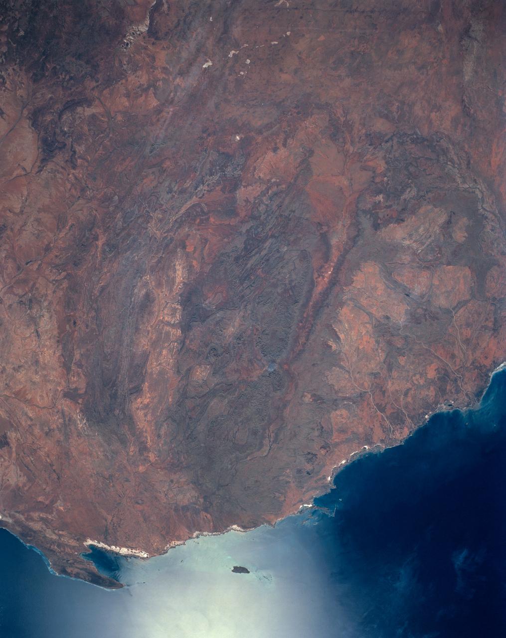 The oval shaped basin of the sedimentary rocks of the Hammersley Range, northern Western Australia (23.0S, 119.0E) dominates the center of this near nadir view. The Fortescue River is the remarkably straight, fault controlled feature bordering the Hammersley on the north. Sand dunes are the main surface features in the northeast and southwest. Many dry lakebeds can be seen to the east as light grey colored patches along the watercourses.