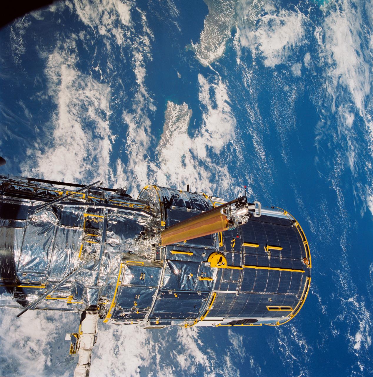 During STS-31, the Hubble Space Telescope (HST) grappled by the remote manipulator system (RMS) end effector is held in appendage deploy position above Discovery, Orbiter Vehicle (OV) 103. The solar array (SA) bistem cassette has been released from its latch fittings. The bistem spreader bars begin to unfurl the SA wing. The secondary deployment mechanism (SDM) handle is visible at the SA end. Stowed against either side of the HST System Support Module (SSM) forward shell are the high-gain antennae (HGA). Puerto Rico and the Dominican Republic are recognizable at the left of the frame.