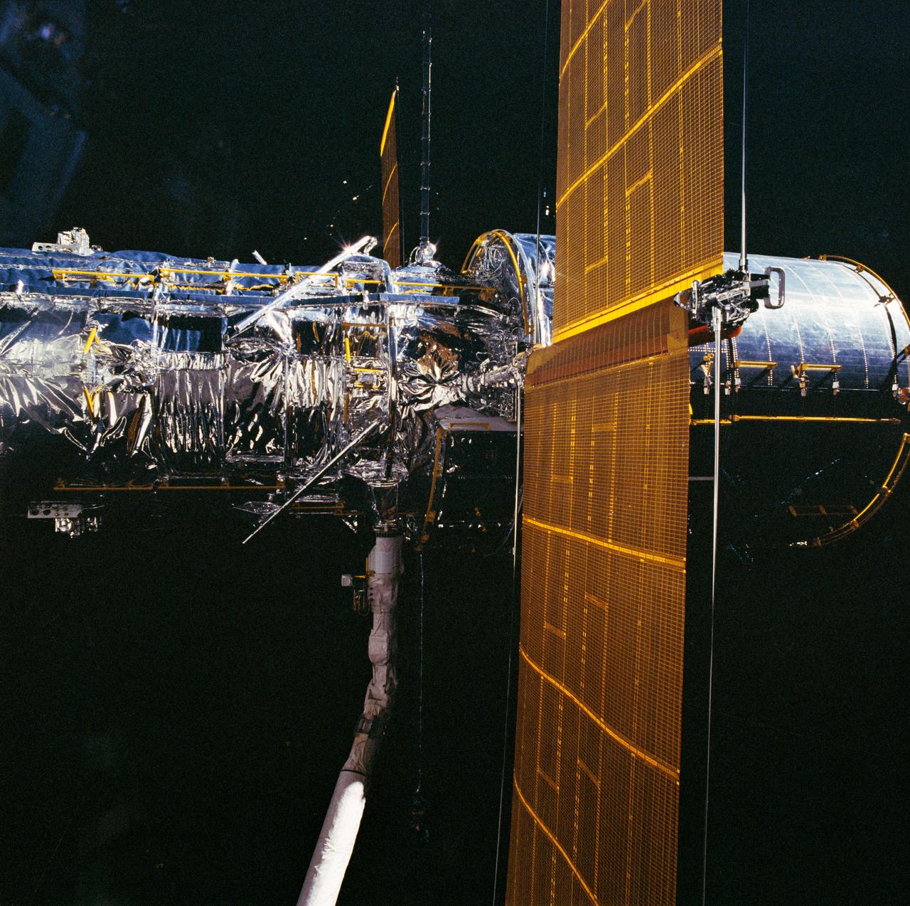During STS-31, the Hubble Space Telescope (HST), grappled by the remote manipulator system (RMS) end effector, is held against the blackness of space. The two solar array (SA) wings (large gold panels) are fully extended with bistem cassette and secondary deployment mechanism (SDM) handle clearly visible. The two deployed high gain antennae (HGA) masts are parallel to the SA panels. RMS end effector is positioned on the starboard fixture during the predeployment checkout operations above Discovery's, Orbiter Vehicle (OV) 103's, payload bay (PLB).