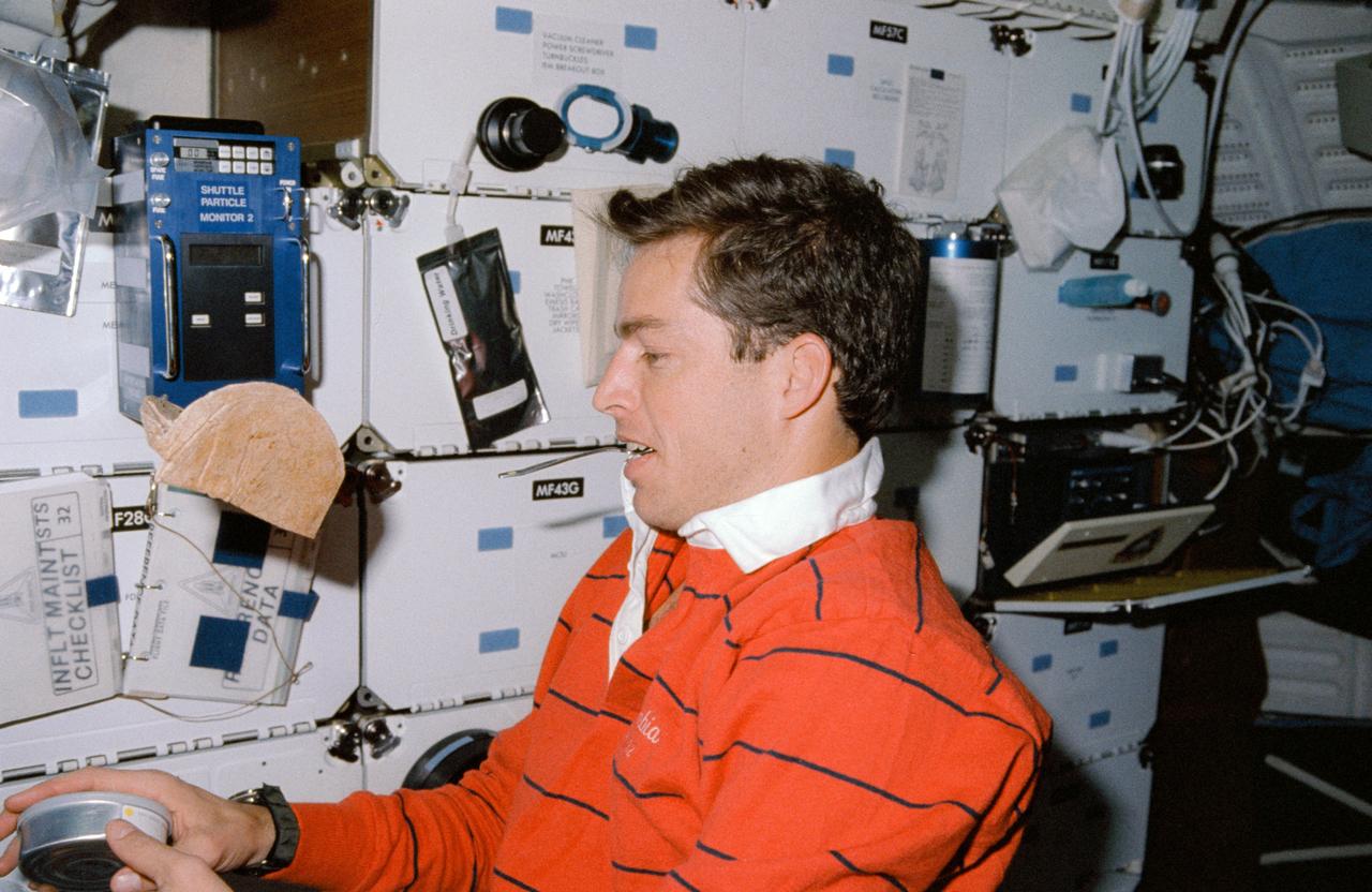 STS032-03-026 (9 - 20 Jan 1990)  --- Astronaut James D. Wetherbee, pilot, having run out of shelf space, uses his mouth to temporarily "park" a spoon during meal time preparations on the mid-deck of the Earth-orbiting Space Shuttle Columbia.  He prepares to open a can while a packet of tortillas floats in the foreground.  This picture was used by the astronauts at their January 30, 1990 Post Flight Press Conference (PFPC) at Johnson Space Center (JSC).