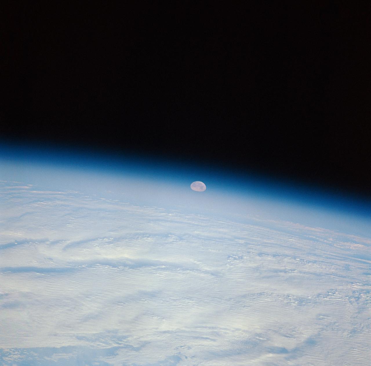 STS-32 crew took this view of the moon setting over the Earth's limb. Near the center is a semi-vortex in the clouds - a storm system in the early stages of formation. The moon's image is distorted due to refraction through the Earth's atmosphere. The near side of the moon is visible showing the vast area of the moon's western seas (Mare Occidental), Apollo landing sites: Apollo 14 at Fra Mauro and Apollo 16 at Central Highlands near Descartes.