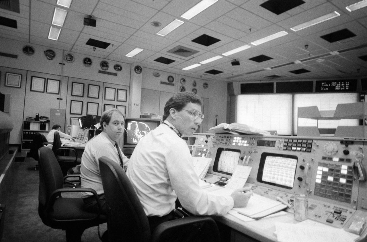 Flight directors Robert E. Castle, Jr (left) and Ronald D. Dittemore monitor console displays during STS-34 mission. Castle and Dittemore will oversee flight activities from their stations in JSC's Mission Control Center (MCC) Bldg 30 flight control room (FCR). In the background are the FCR large screens which display flight data.