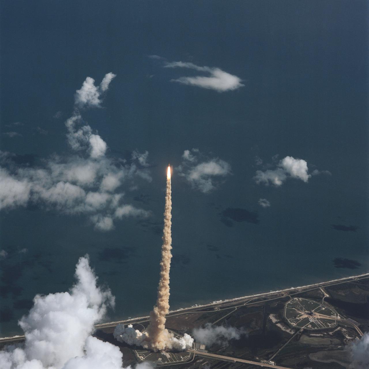 STS-34 Atlantis, Orbiter Vehicle (OV) 104, lifts off from Kennedy Space Center (KSC) Launch Complex (LC) Pad 39B at 12:53:39:983 pm Eastern Daylight Time (EDT). This aerial view shows OV-104, its external tank (ET), and two solid rocket boosters (SRBs) rising high above LC Pad 39B atop a plume of exhaust smoke. Atlantic Ocean is visible in the background. The liftoff marks the beginning of a five-day mission in space.