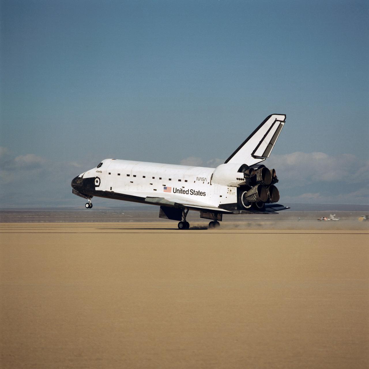 STS-34 Atlantis, Orbiter Vehicle (OV) 104, main landing gear (MLG) touches down on Runway 23 dry lake bed at Edwards Air Force Base (EAFB), California. The nose landing gear rides above runway before touchdown as the MLG wheels produce a cloud of dust. OV-104's port side profile is captured as it glides by at a speed of approximately 195 knots (224 miles per hour). The tail section with deployed speedbrake/rudder and space shuttle main engines (SSMEs) are visible.