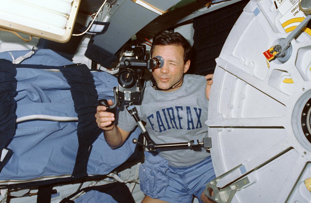 STS-36 Mission Specialist (MS) Pierre J. Thuot operates 16mm ARRIFLEX motion picture camera mounted on the open airlock hatch via a bracket. Thuot uses the camera to record activity of his fellow STS-36 crewmembers on the middeck of Atlantis, Orbiter Vehicle (OV) 104. Positioned between the airlock hatch and the starboard wall-mounted sleep restraints, Thuot, wearing a FAIRFAX t-shirt, squints into the cameras eye piece. Thuot and four other astronauts spent four days, 10 hours and 19 minutes aboard OV-104 for the Department of Defense (DOD) devoted mission.
