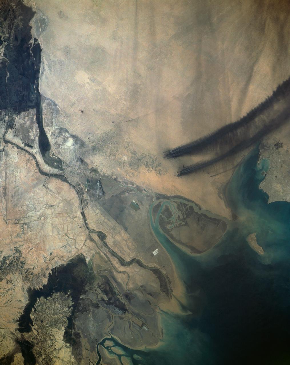 Smoke from the burning oil fields to the north of Kuwait City, seen on the south shore of Kuwayt Bay, almost totally obscures the view of the tiny, but oil rich, nation of Kuwait (30.0N, 48.0E). During the brief war between Iraq and the Allied forces, many of the oil wells in Kuwait were destroyed and set afire. For several months, those fires burned out of control, spewing wind borne smoke and ash for hundreds of miles.