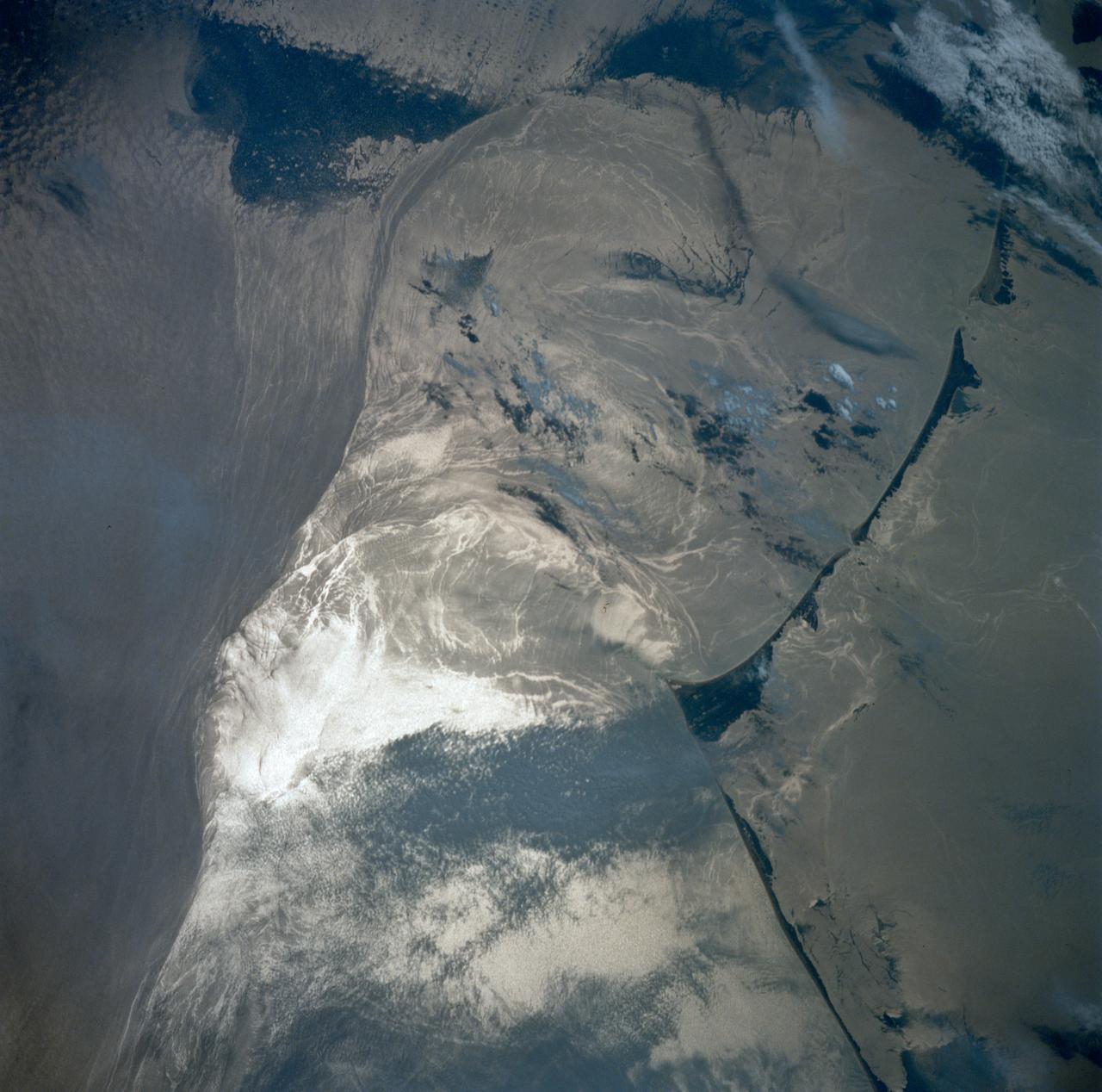 STS039-83-091 (28 April-6 May 1991) --- (HOLD PHOTO WITH BARRIER ISLANDS ALONG THE LEFT SIDE).  This photograph of the water sunglint pattern along the Outer Banks of North Carolina was taken with a 70mm handheld camera equipped with color visible film and a 250mm lens.  The islands appear as dark areas along the left side of the frame and Cape Hatteras is seen to the left and slightly above frame center. Sunglint is composed of light reflected from the water surface and the patterns seen in this photo are largely the result of sea surface roughness. Smooth water has a mirror-like reflectance and is represented in the photo as a bright area.  Rough water, on the other hand, tends to disperse the light reflected from the water surface and, therefore, appears dark.  Water currents in this area are extremely dynamic because the swift, northward flowing Gulf Stream (the offshore darker water along the right side of the photo) diverges from the coastline north of Cape Hatteras.  The sharp line between bright and dark water is the western boundary of the Gulf Stream.