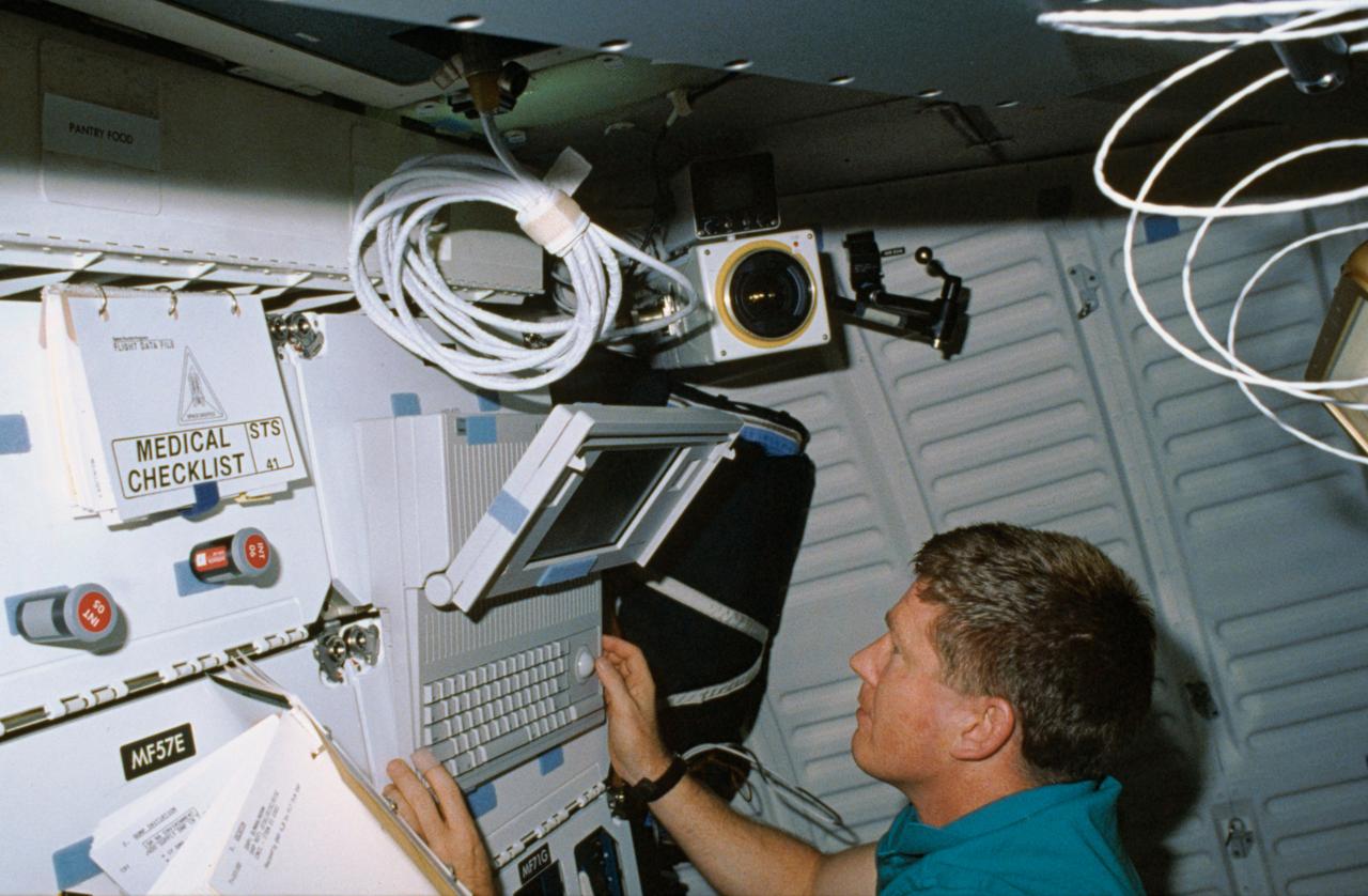 STS-41 Mission Specialist (MS) William M. Shepherd uses Detailed Test Objective (DTO) Space Station Cursor Control Device Evaluation MACINTOSH portable computer on the middeck of Discovery, Orbiter Vehicle (OV) 103. The computer is velcroed to forward lockers MF71C and MF71E. Surrounding Shepherd are checklists, the field sequential (FS) crew cabin camera, and a lighting fixture.