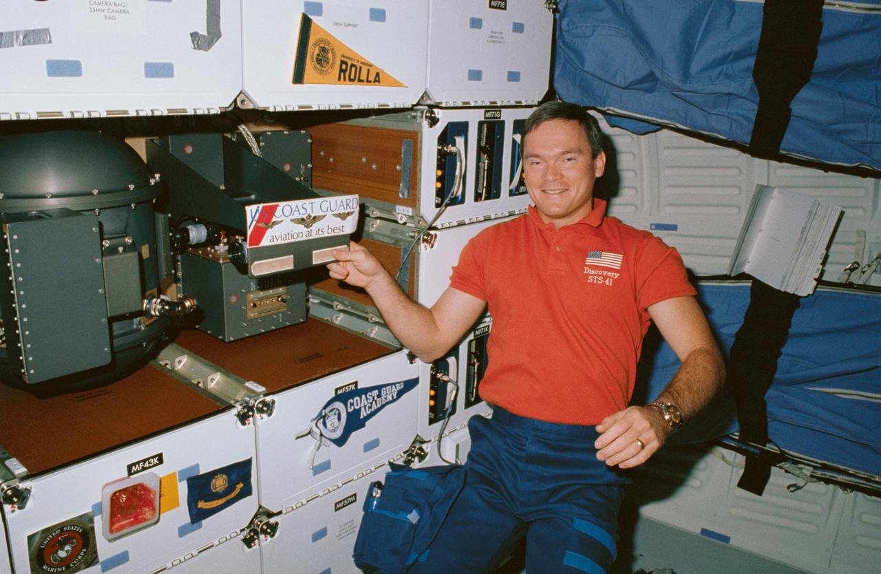 STS041-06-029 (10 Oct 1990) --- STS-41 Mission Specialist (MS) Bruce E. Melnick, who is a graduate of the Coast Guard Academy and the first ever active Coast Guardsman to fly in space, draws attention to his branch of the service, while posing next to a banner from his Alma Mater (on locker MF57K) and a United States (U.S.) Coast Guard decal (on the Solid Surface Combustion Experiment (SSCE)). Behind Melnick are the starboard wall-mounted sleep restraints. Insignias belonging to other crewmembers are displayed on the lockers including University of Missouri Rolla and U.S. Marine Corps.