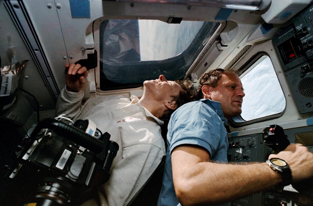 STS-42 Payload Specialist Roberta L. Bondar (light shirt) smiles as she looks out overhead window W7 while Mission Specialist (MS) and Payload Commander (PLC) Norman E. Thagard peers out aft viewing window W9. The two crewmembers are on the aft flight deck of Discovery, Orbiter Vehicle (OV) 103. In the foreground, a camera is ready to capture the Earth below.