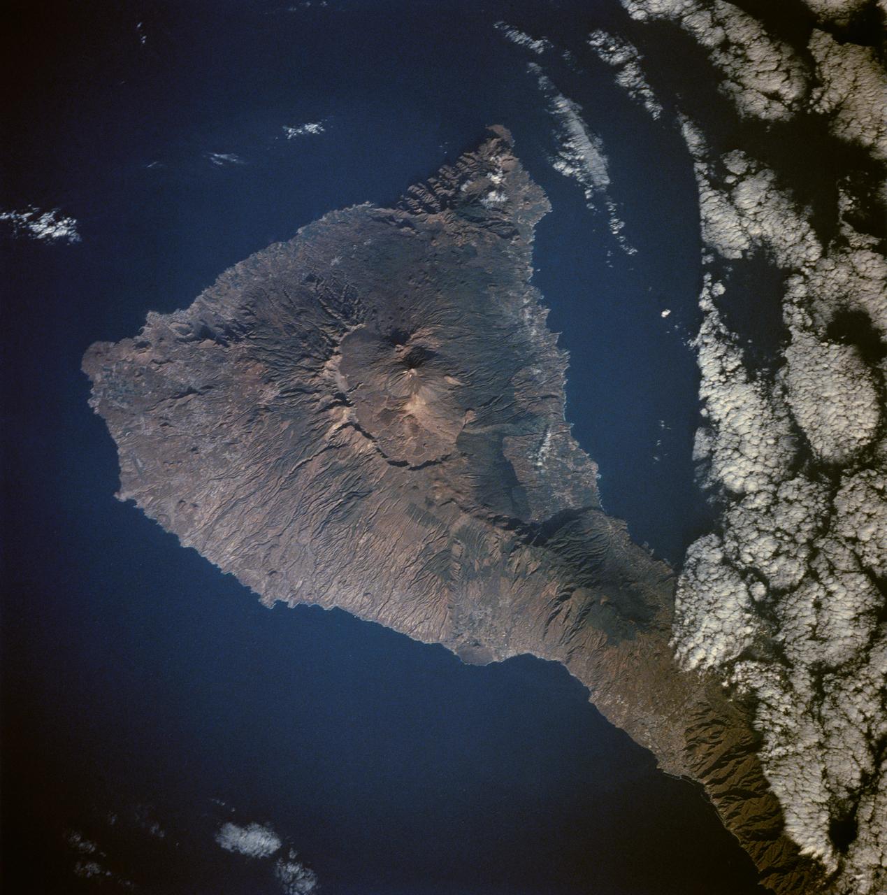 Tenarife Island is one of the most volcanically active of the Canary Island archipelago, Atlantic Ocean, just off the NW coast of Africa, (28.5N, 16.5W). The old central caldera, nearly filled in by successive volcanic activity culminating in two stratocones. From those two peaks, a line of smaller cinder cones extend to the point of the island. Extensive gullies dissect the west side of the island and some forests still remain on the east side.