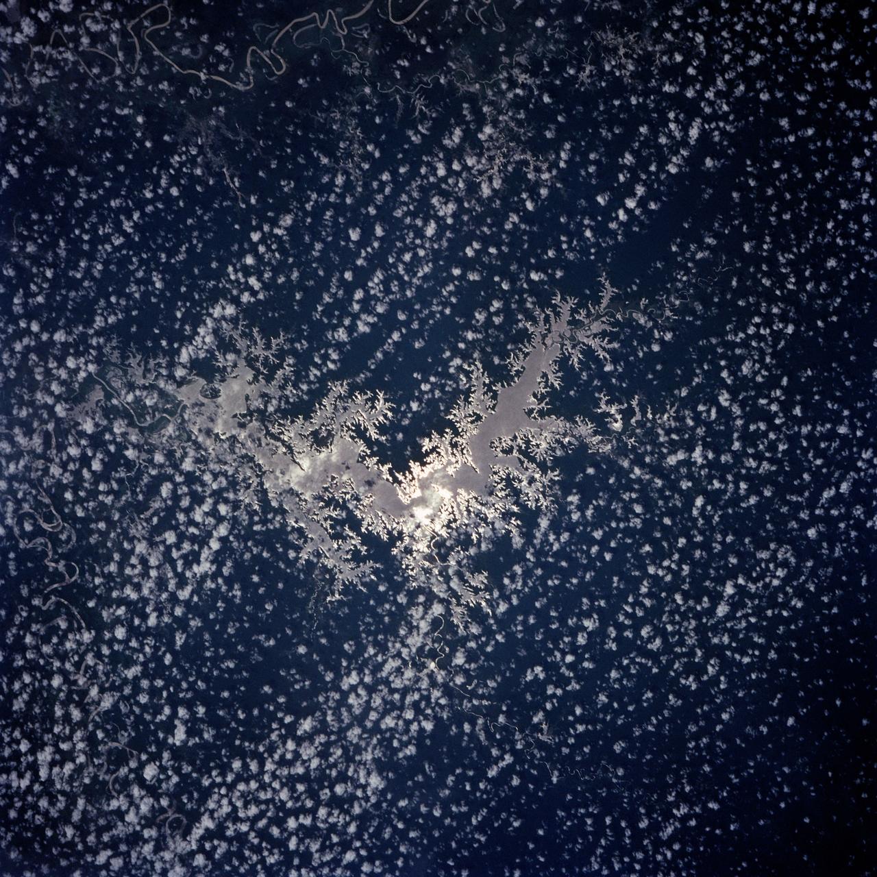 Lake Murray, a manmade reservoir, lies between the Fly and Strickland River Basins, Papua, New Guinea (7.0S, 141.5E). The region, photographed in sunglint, shows the water level in the reservoir and the full extent of the drainage basins of both river systems as the rivers meander through wide alluvial floodplains. Some forest clearing can be seen in places throughout the region, but most of the area remains in closed canopy forest.