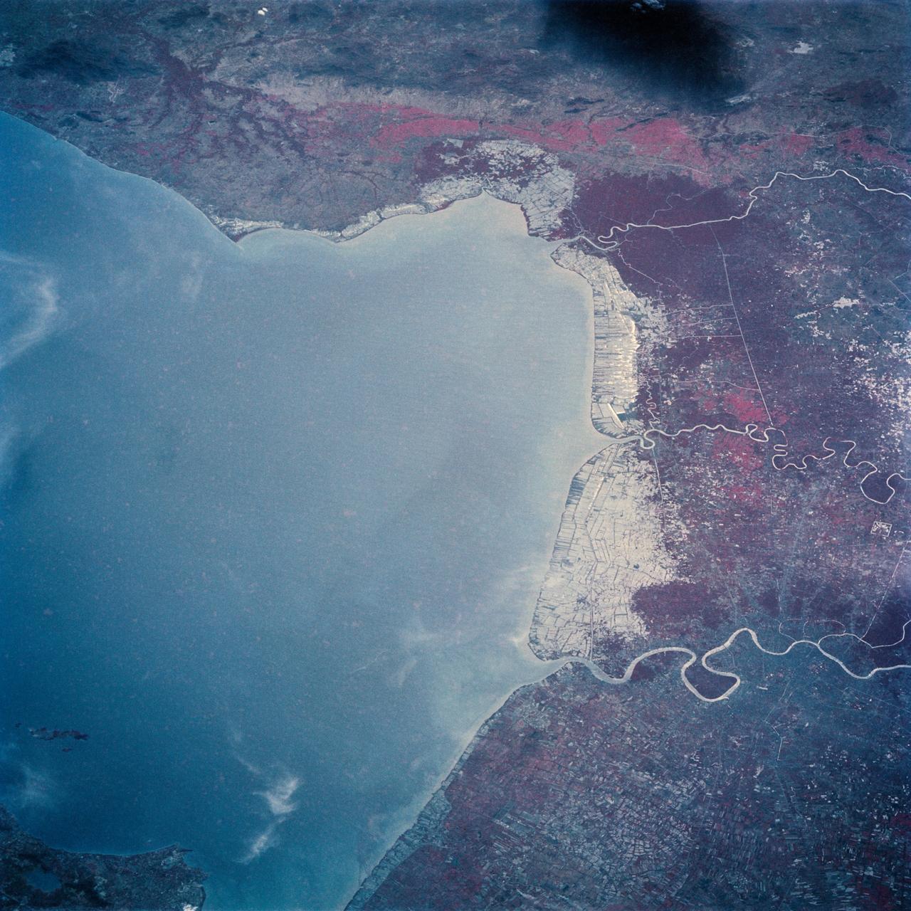 The Bight of Bangkok and the city of Bangkok, Thailand are visible in this west looking view. The city of Bangkok, with an estimated population in excess of 3.7 million people, can be seen in the lower right-hand corner of the view. The city, famous as a jewelry trading center, dealing in precious stones and silver and bronze ware, is situated in a vast lowland. This lowland is a major rice growing area interspersed with canals used for irrigation and drainage. Rice is a major export commodity of Thailand. The deforested hills of the Bilauktaung Range can be seen at the top of the view.