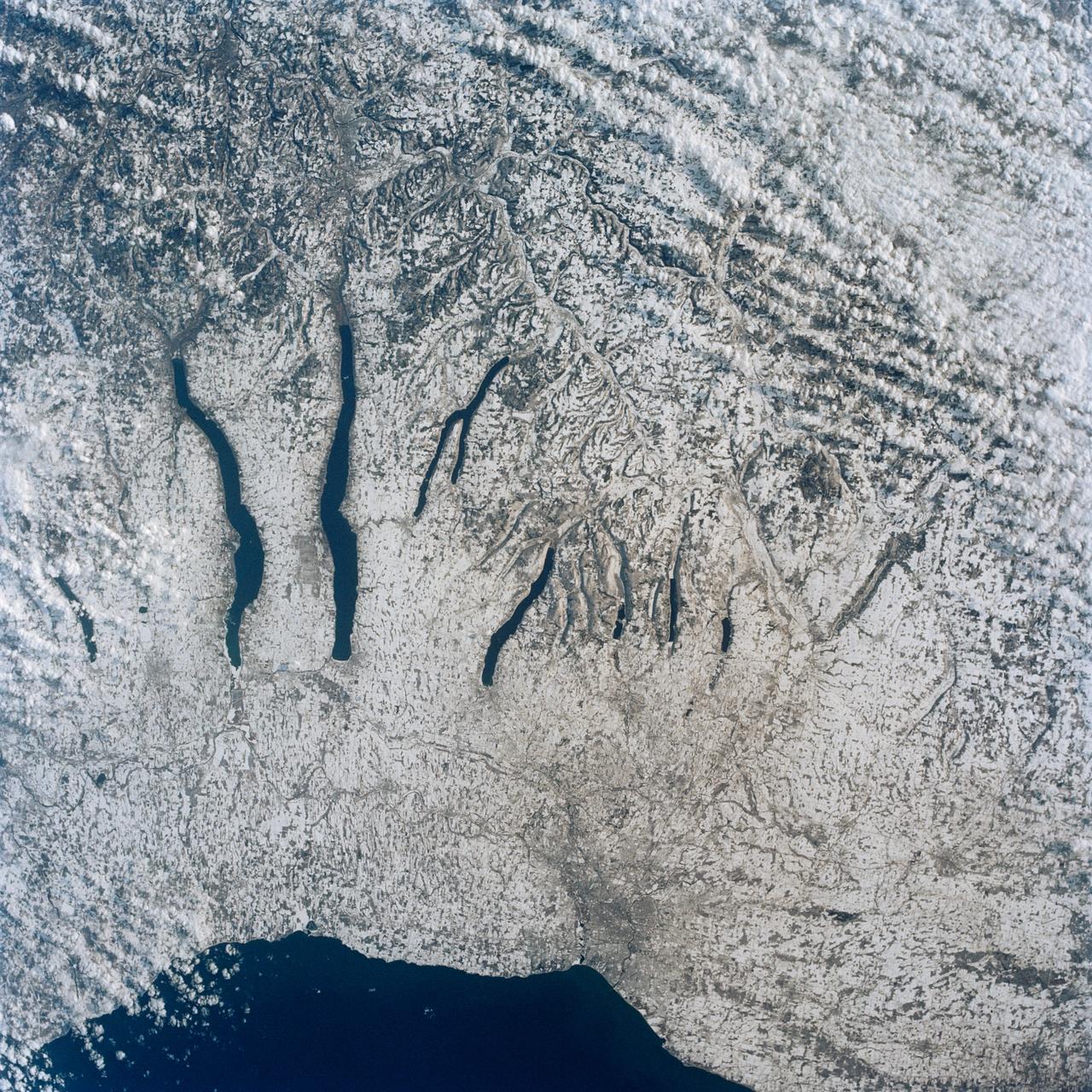 This view of the central portion of upstate New York, centers on the Finger Lakes. The large city on the shore of Lake Ontario, is Rochester. Although the city, being a business, educational and technical center, has no heavy industry, the outline of the city shows fairly well in the snow, but not as well as the outlines of industrial cities elsewhere in the world. The Finger Lakes are large linear lakes carved out by glaciers during the last ice age.