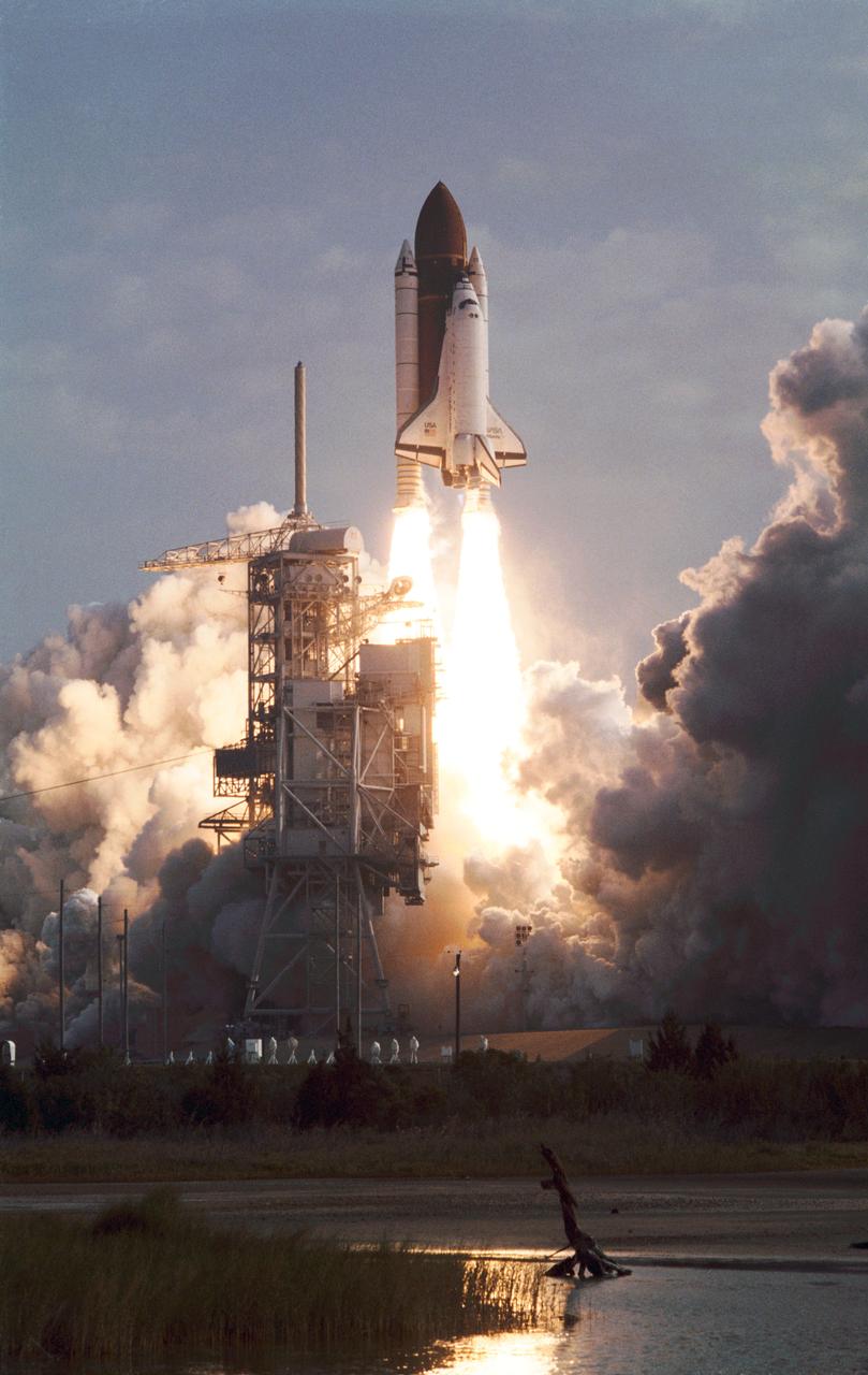 STS-45 Atlantis, Orbiter Vehicle (OV) 104, lifts off from a Kennedy Space Center (KSC) Launch Complex (LC) Pad at 8:13:40:048 am (Eastern Standard Time (EST)). Exhaust billows out the solid rocket boosters (SRBs) as OV-104 atop its external tank (ET) soars above the mobile launcher platform and is nearly clear of the fixed service structure (FSS) tower. The diamond shock effect produced by the space shuttle main engines (SSMEs) is visible. The glow of the SRB/SSME firings is reflected in a nearby waterway. An exhaust cloud covers the launch pad area.