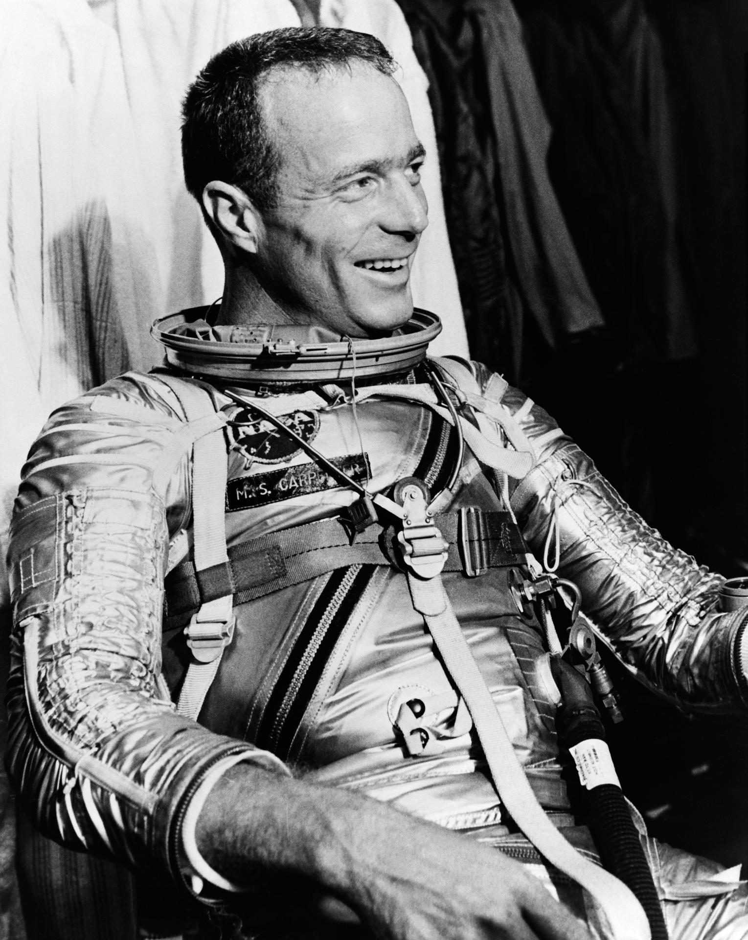 Astronaut Scott Carpenter smiles as he sits in a chair wearing his silver space suit.