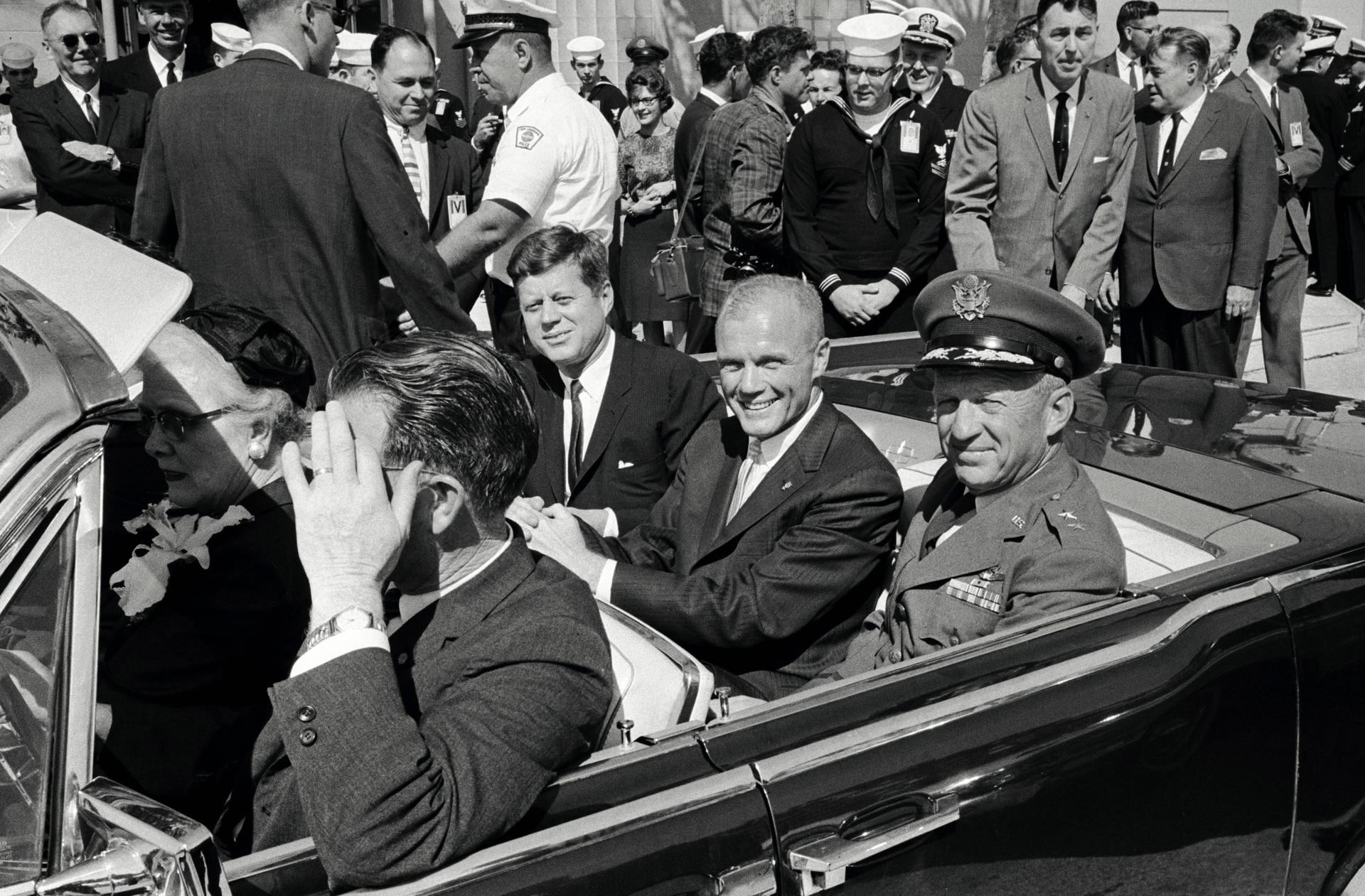 President John F. Kennedy is seated next to John Glenn in a car during a parade.