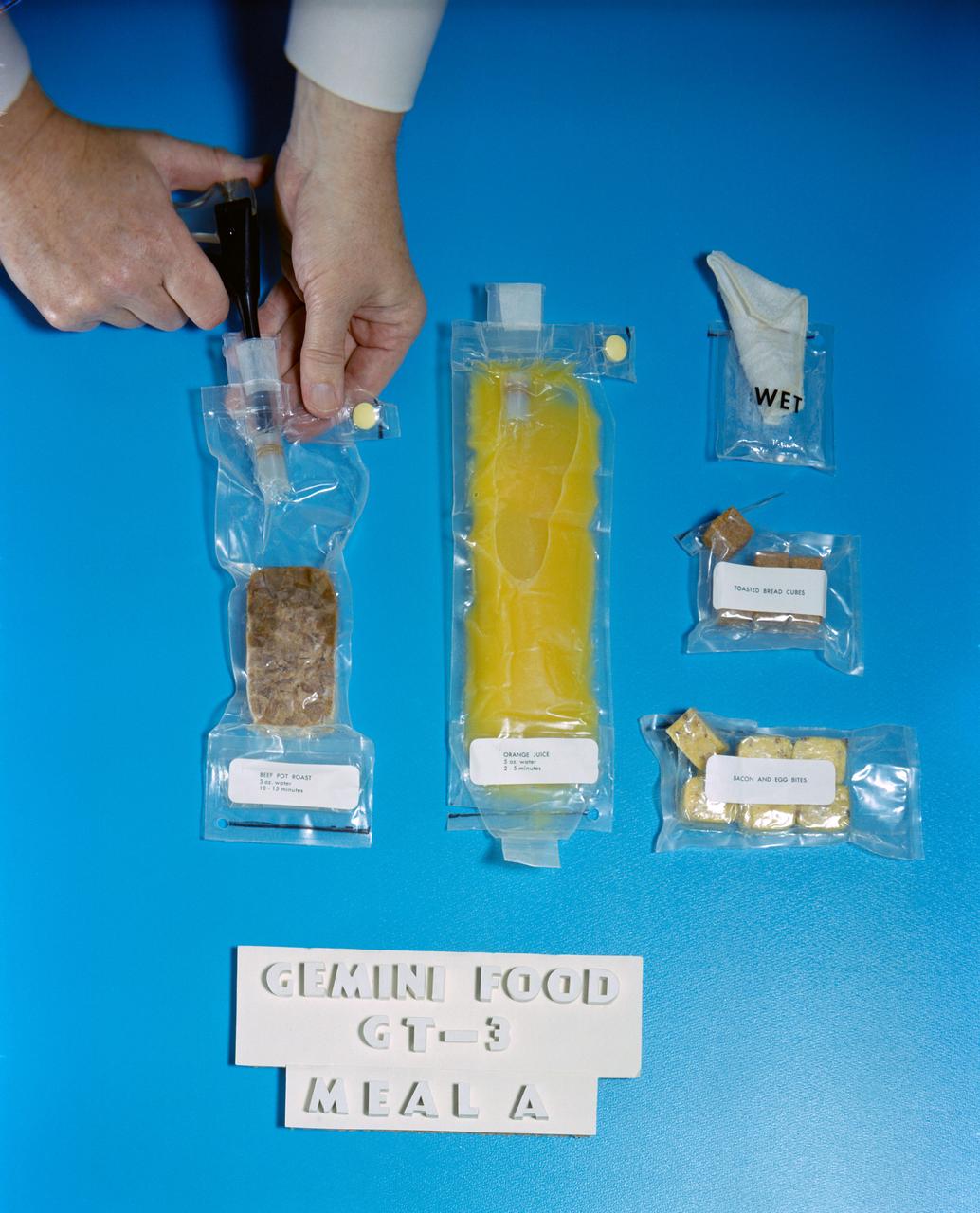 S65-10971 (March 1965 ) --- Food packets for use on the Gemini-3 flight including dehydrated beef pot roast, bacon and egg bites, toasted bread cubes, orange juice and a wet wipe. Water is being inserted into the pouch of dehydrated food.
