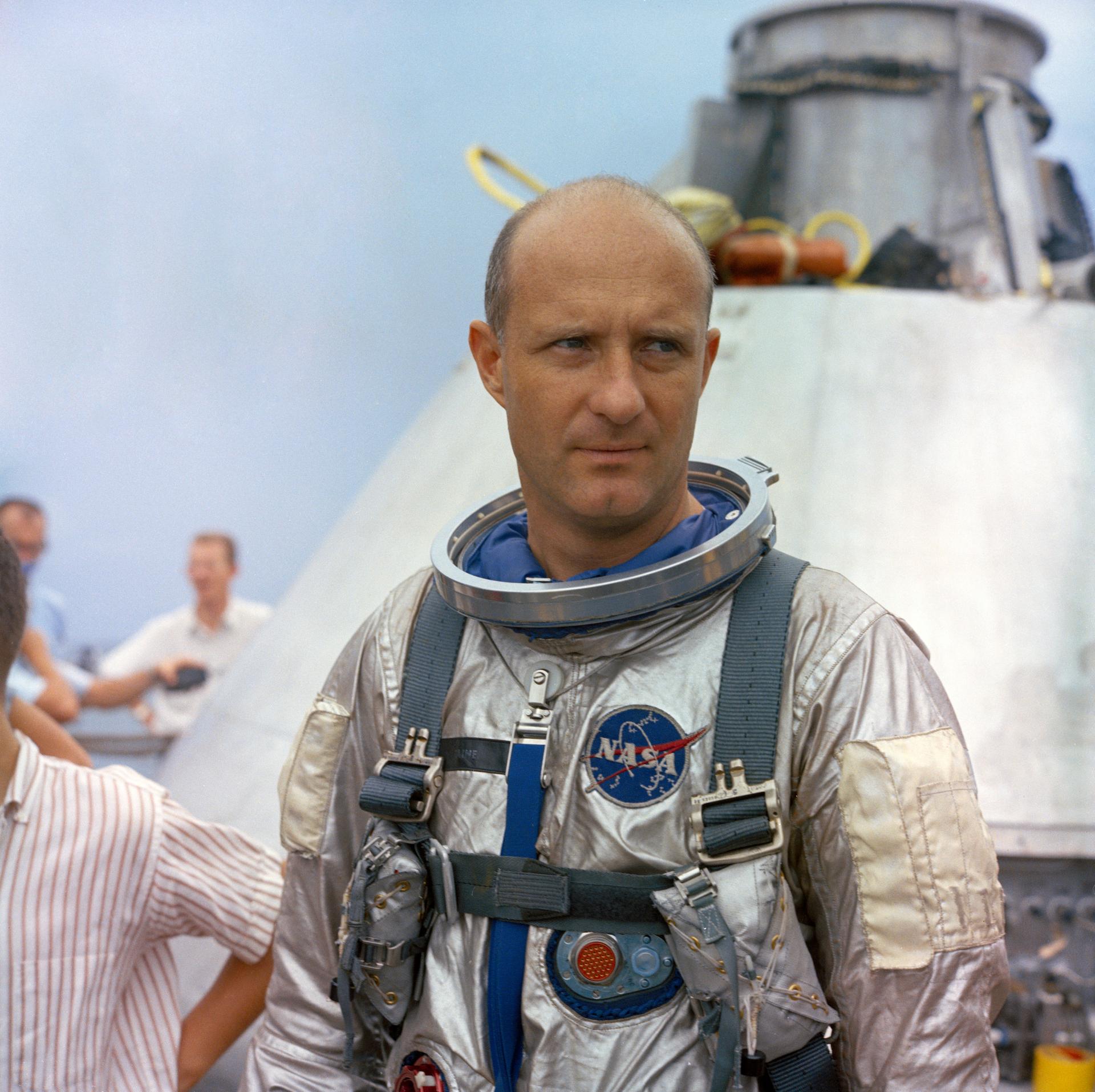 An astronaut in a silver spacesuit standing in front of an Apollo space capsule.