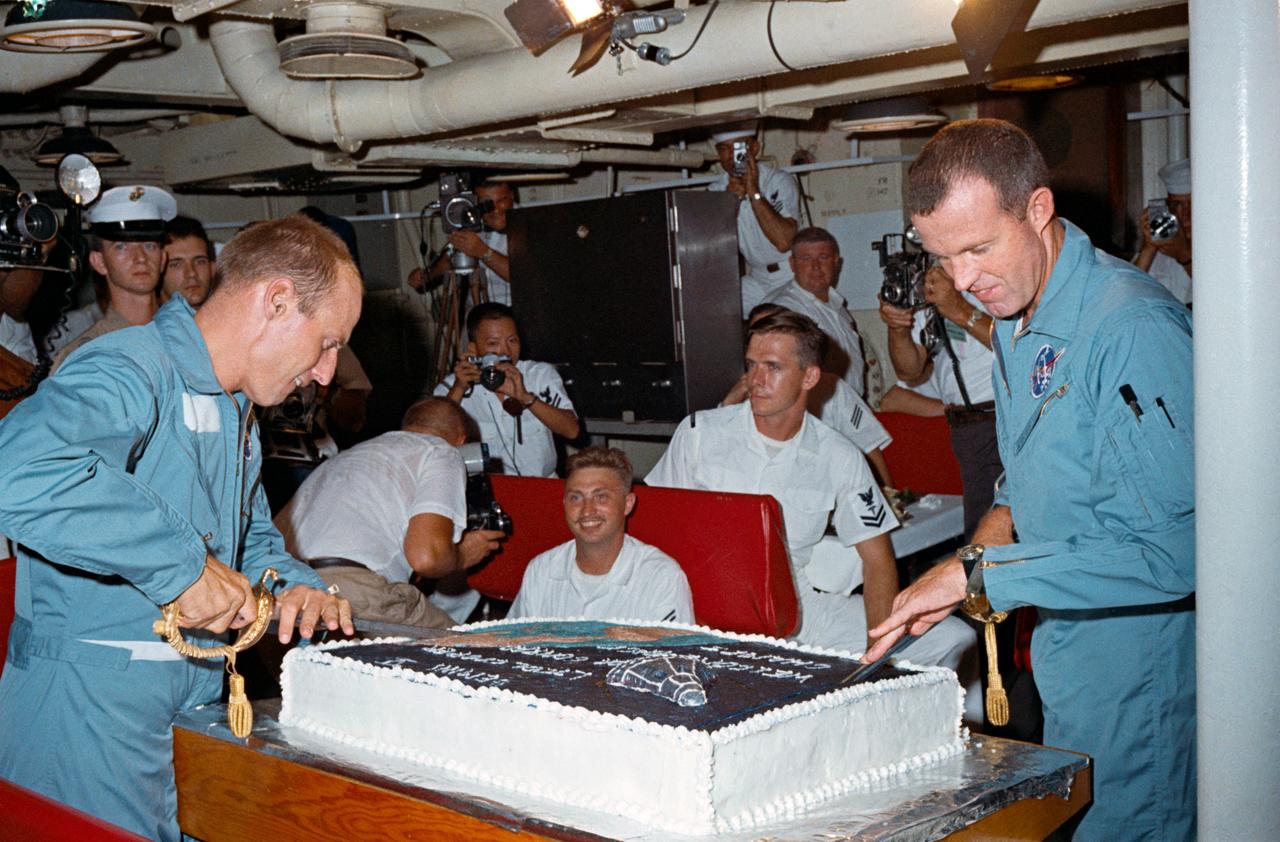S65-51660 (29 Aug. 1965) --- Astronauts Charles Conrad Jr. (left) and L. Gordon Cooper Jr. prepare to slice into the huge cake prepared for them by the cooks onboard the aircraft carrier USS Lake Champlain. They are using ornamental Navy swords for knives.