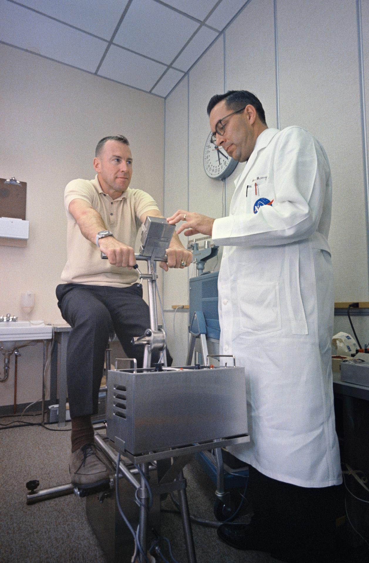 Astronaut Jim Lovell sits on an exercise bike while Dr. Charles Berry checks on him