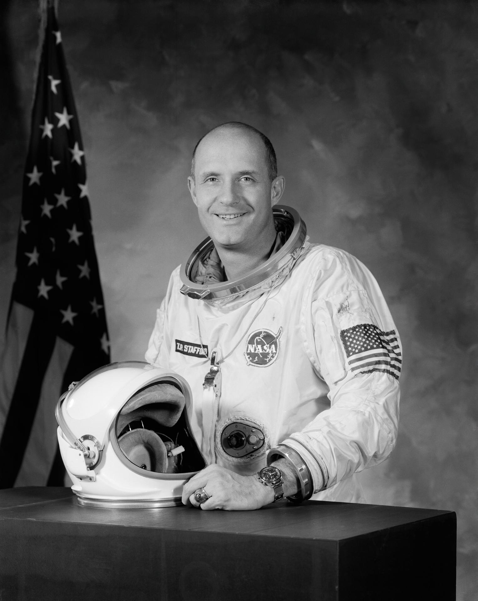 Black and white portrait of Thomas P. Stafford in white spacesuit in front of US flag