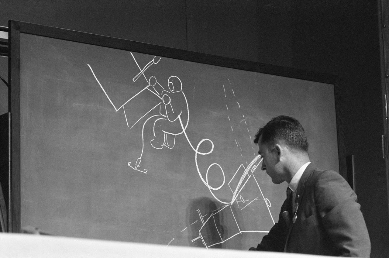 S66-39897 (1 Aug. 1966) --- Astronaut John W. Young, Gemini-10 command pilot, uses a chalk drawing on a blackboard to illustrate how astronaut Michael Collins, Gemini-10 pilot, looked when he inspected the Agena Target Docking Vehicle during his extravehicular activity. Young was discussing the mission before a gathering of news media representatives in the Building 1 auditorium. Photo credit: NASA
