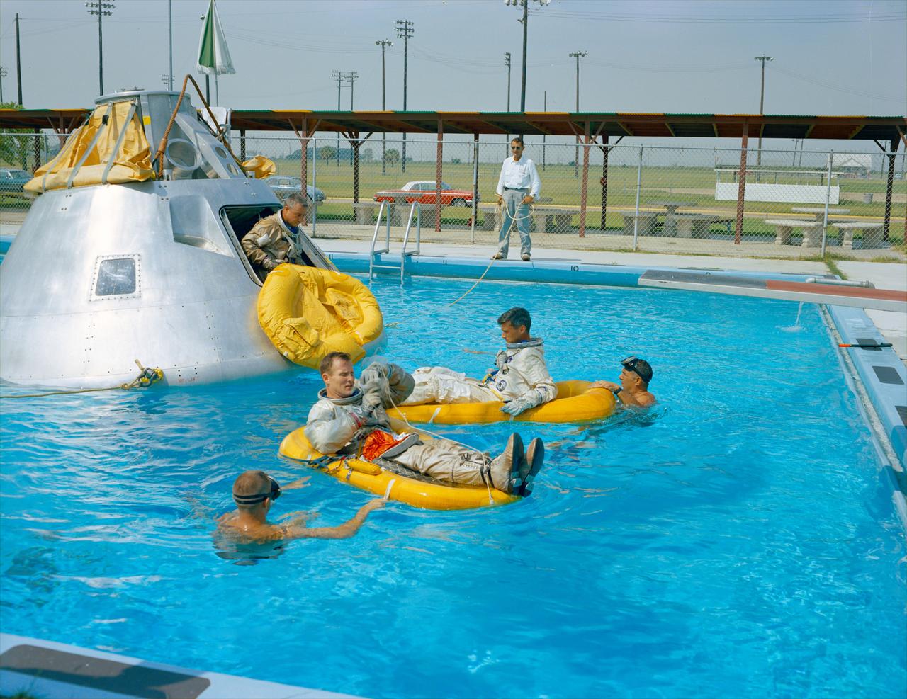 Boilerplate (B/P) model of the Apollo Spacecraft resting nose down in the swimming pool at EAFB during a training session with the first (1st) crew named by NASA. NASA swimmers are in the water to assist during the practice session. Inside the spacecraft are Astronauts  Roger B. Chaffee, and Edward H. White II, members of the crew. Astronaut Virgial I. Grissom is visible in the entrance to the craft.
