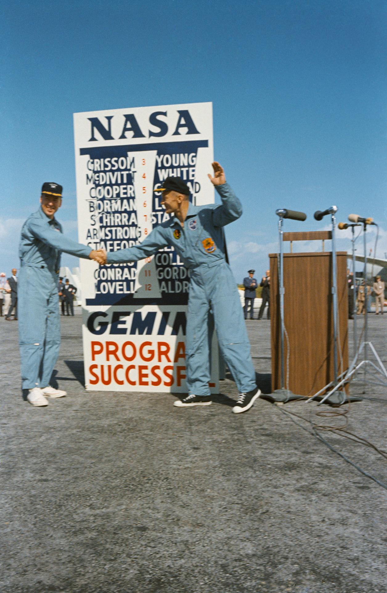 Jim Lovell and Buzz Aldrin shake hands in front of a sign celebrating the successes of the Gemini Program