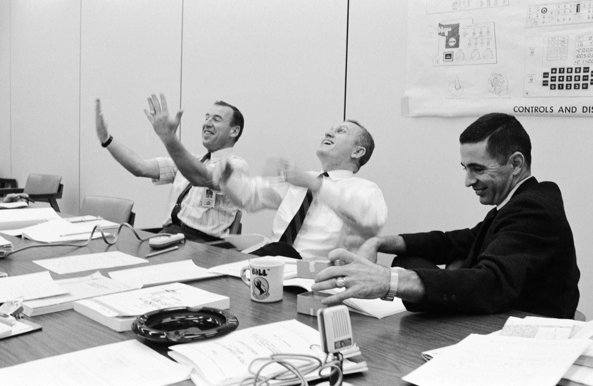 The Apollo 8 crew sits at a table during a debriefing. Lovell and Borman are gesturing dramatically while Anders smiles.