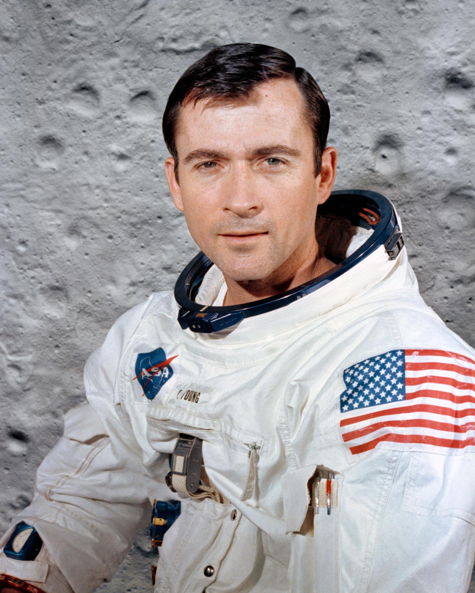 Portrait of John Young. He wears a white space suit and is posed in front of a backdrop of the Moon's surface.