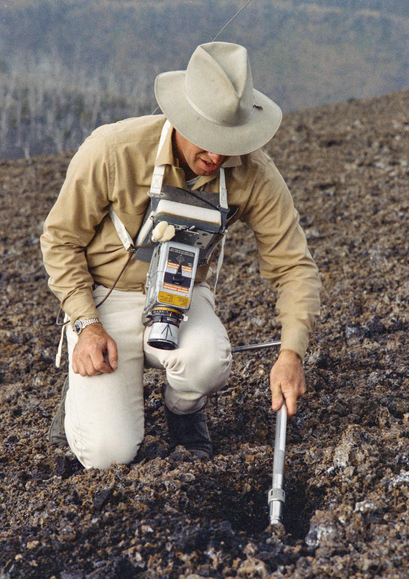 Jim Lovell crouches down on a rocky surface with a bulky camera strapped to his chest.