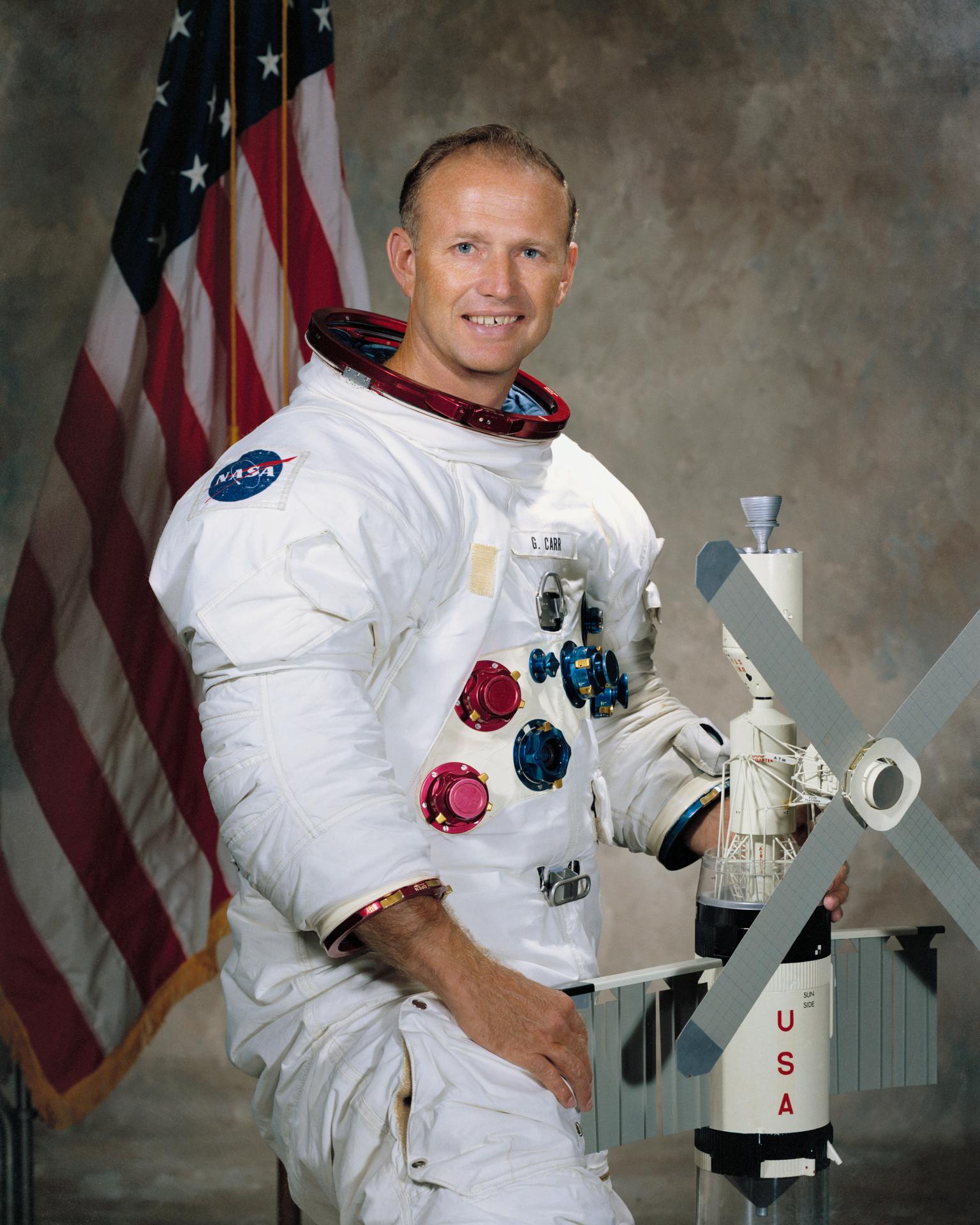 A man stands in a spacesuit with his helmet off, holding a model of Skylab 4