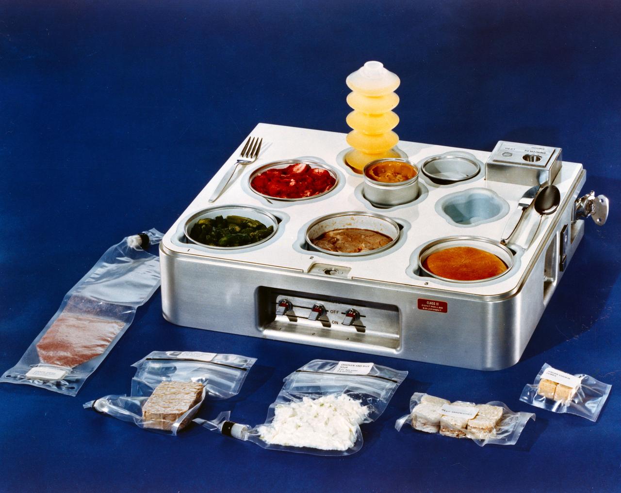 S72-15409 (1972) --- A close-up view of a food tray which is scheduled to be used in the Skylab program. Several packages of space food lie beside the tray. The food in the tray is ready to eat. Out of tray, starting from bottom left: grape drink, beef pot roast, chicken and rice, beef sandwiches and sugar cookie cubes. In tray, from back left: orange drink, strawberries, asparagus, prime rib, dinner roll and butterscotch pudding in the center. Photo credit: NASA