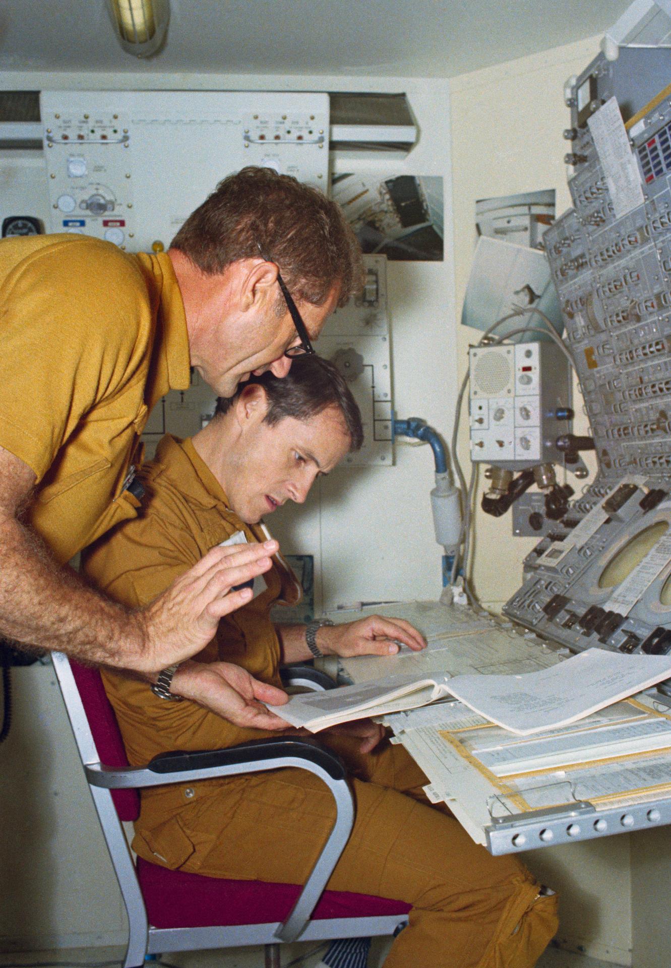 Edward Gibson is seated in a chair while William Pogue leans over him as they discuss procedures at the Apollo Telescope Mount mockup