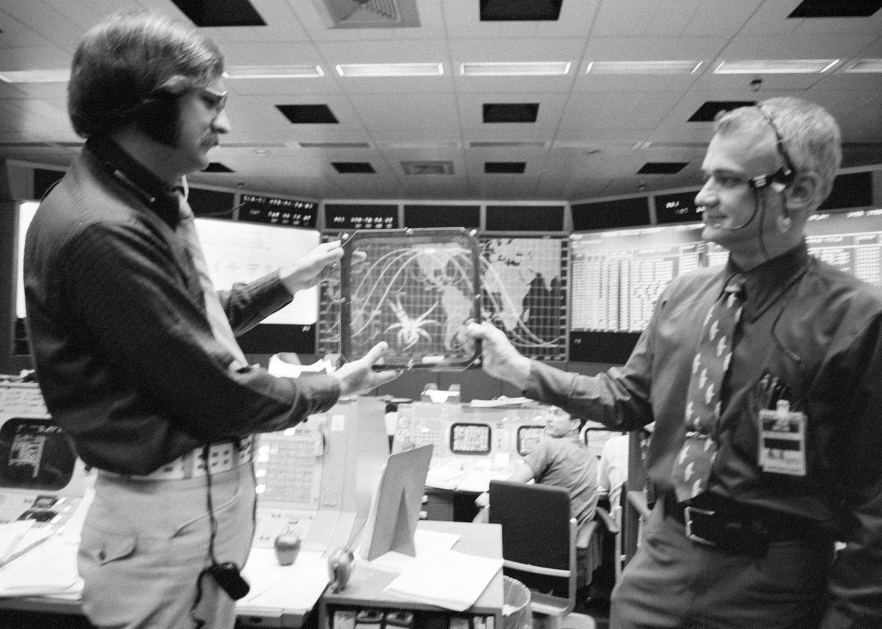 S73-34456 (September 1973) --- Flight Director Neil B. Hutchinson, left, and astronaut Bruce McCandless II hold up a glass enclosure - home for the spider Arachne, which is the same species as the two spiders carried on the Skylab 3 mission. The real spider is the one barely visible at the upper right corner of the square; the larger one is a projected image on the rear-screen-projected map in the front of the Mission Operations Control Room (MOCR) of the Mission Control Center (MCC). McCandless served as backup pilot for the first manned Skylab mission and was a spacecraft communicater (CAPCOM) for the second crew. Photo credit: NASA