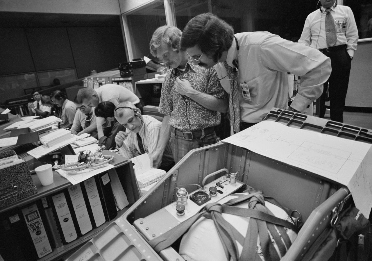 S73-37030 (November 1973) --- The procedures for repairing the coolant system aboard the Airlock Module of the Skylab space station in Earth orbit are discussed by flight controllers in the Mission Operations Control Room in the Mission Control Center at Johnson Space Center. Skylab 4 flight director Neil Hutchinson is on the right. Astronaut Russell L. Schweickart is wearing the sports shirt. Astronaut Bruce McCandless II, a Skylab 4 CAPCOM, is seated next to Schweickart. Items of equipment in the foreground are similar to components of a special coolant re-servicing kit which was taken to Earth orbit by the Skylab 4 crewmen. The kit consists of a tank containing 42 pounds of COOLANOL, a series of saddle valves, bolts and spacers, and leak-check hoses. The re-supply tank is a modified command module reaction control subsystem (RCS) fuel tank. Photo credit: NASA