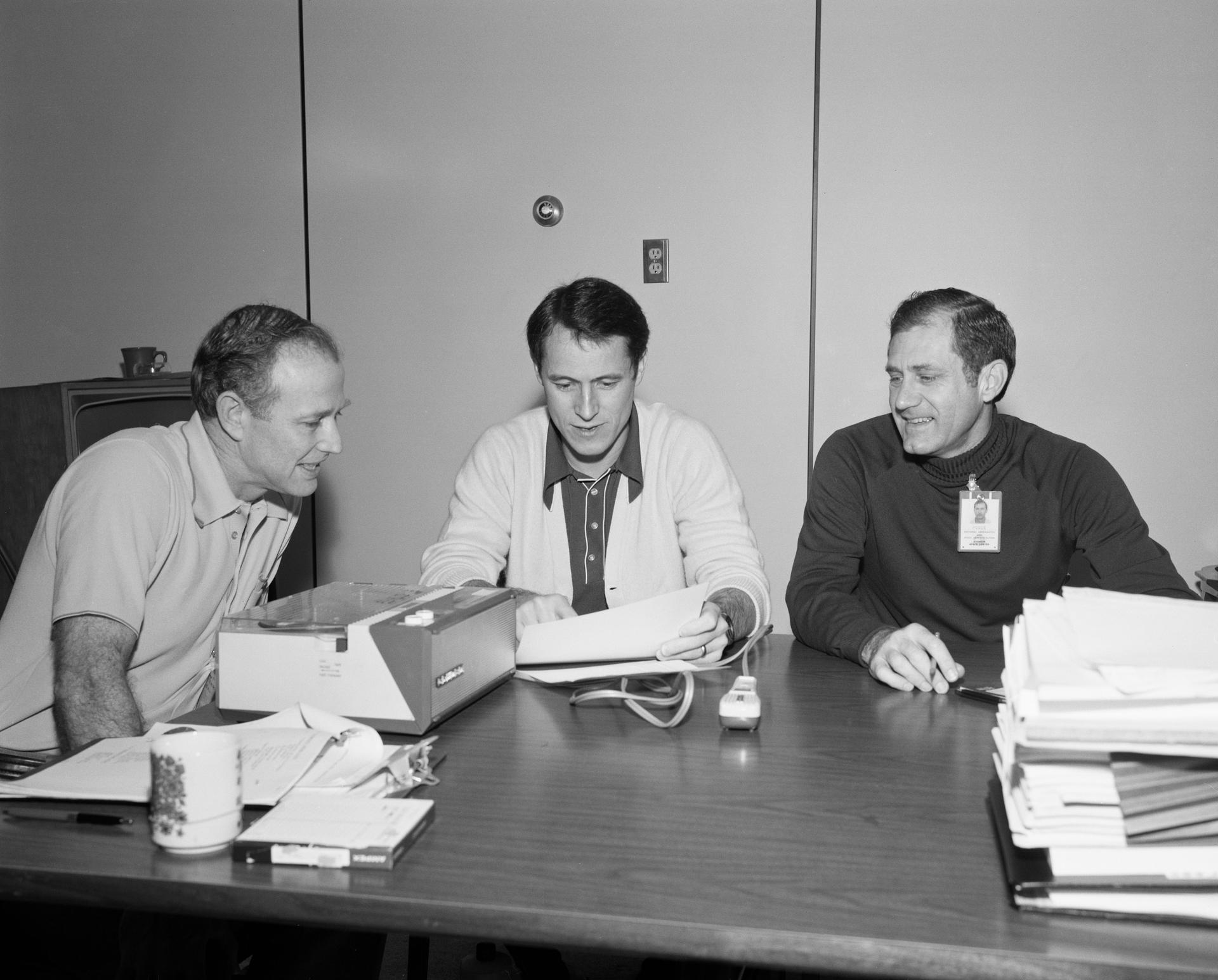 The three crewmen for Skylab 4 are seated at a table, looking over their notes for their upcoming post-mission press conference.