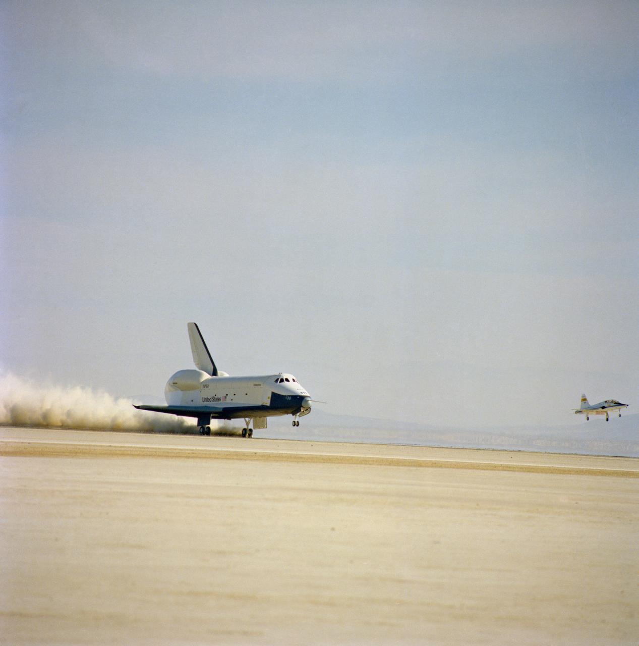 S77-28144 (13 Sept 1977) --- The shuttle orbiter 101 "Enterprise" stirs up a cloud of desert sand and dust as it lands to conclude a five-minute, 28-second unpowered flight during the second free flight of the Shuttle Approach and Landing Tests (ALT) conduced on September 13, 1977 at the Dryden Flight Research Center in Southern California. The Orbiter 101 crew astronauts Joe H. Engle, commander, and Richard H. Truly, pilot. The ALT free flights are designed to verify orbiter subsonic airworthiness, integrated systems operations and pilot-guided approach and landing capability and satisfy prerequisites to automatic flight control and navigation mode. On the right a T-38 chase plane comes in for a landing, also. This picture was taken with an Air Force Test Center 70mm Hasselblad camera installed on a tracking mount parked on the lake bed.