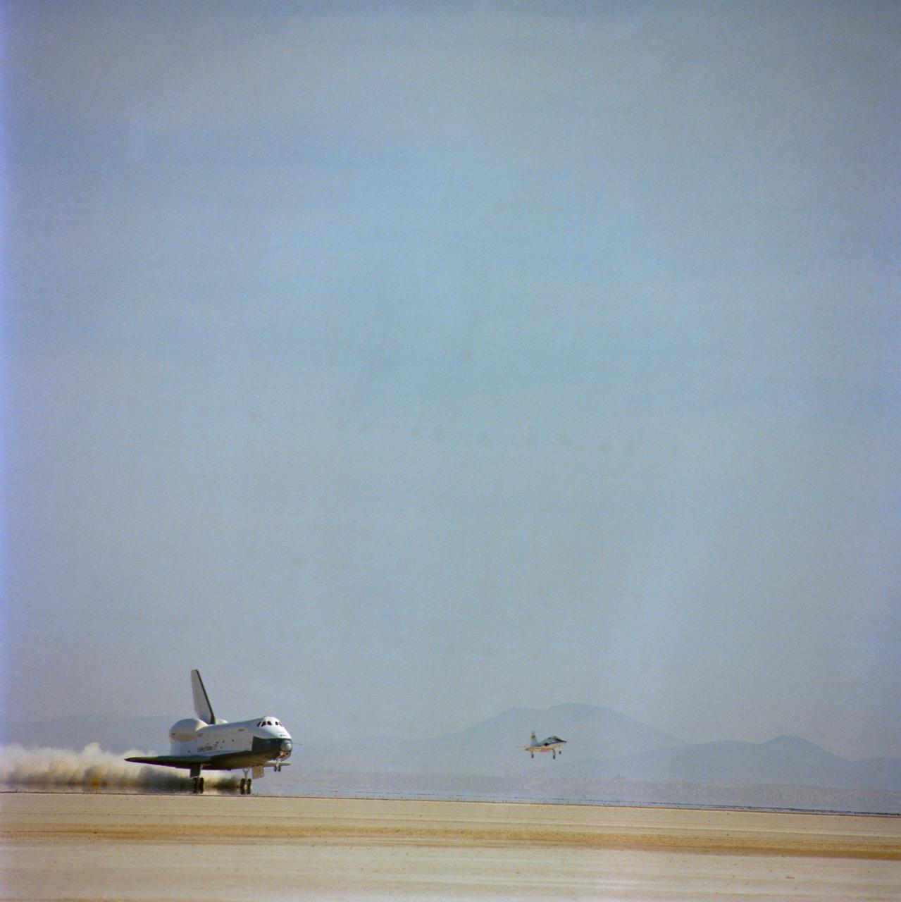 S77-28327 (13 Sept 1977) --- The shuttle orbiter 101 "Enterprise" stirs up a cloud of desert sand and dust as it lands to conclude a five-minute, 28-second unpowered flight during the second free flight of the Shuttle Approach and Landing Tests (ALT) conduced on September 13, 1977 at the Dryden Flight Research Center in Southern California. The Orbiter 101 crew astronauts Joe H. Engle, commander, and Richard H. Truly, pilot. The ALT free flights are designed to verify orbiter subsonic airworthiness, integrated systems operations and pilot-guided approach and landing capability and satisfy prerequisites to automatic flight control and navigation mode. On the right a T-38 chase plane comes in for a landing, also. This picture was taken with an Air Force Test Center 70mm Hasselblad camera installed on a tracking mount parked on the lake bed.