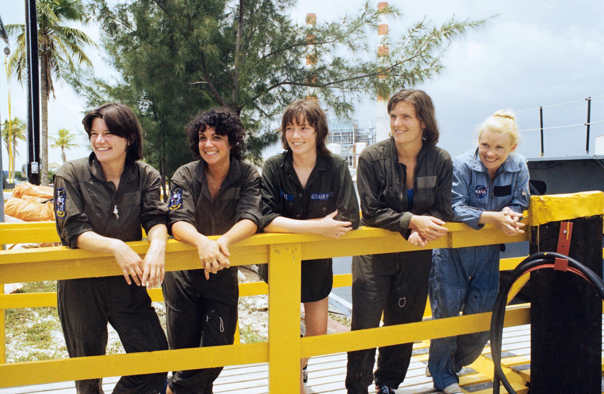 The first five women astronauts leaning up against a fence in green overalls.