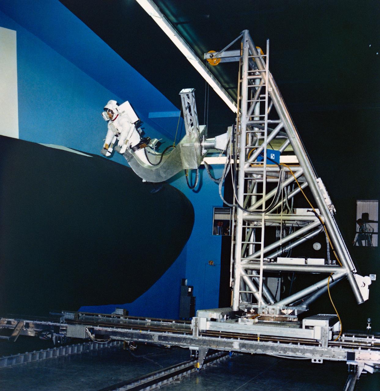 S80-36889 (24 July 1980) ---  Astronaut Bruce McCandless II uses a simulator at Martin Marietta?s space center near Denver to develop flight techniques for a backpack propulsion unit that will be used on Space Shuttle flights.  The manned maneuvering unit (MMU) training simulator allows astronauts to "fly missions" against a full scale mockup of a portion of the orbiter vehicle.  Controls of the simulator are like those of the actual MMU.  Manipulating them allows the astronaut to move in three straight-line directions and in pitch, yaw and roll.  One possible application of the MMU is for an extravehicular activity chore to repair damaged tiles on the vehicle.  McCandless is wearing an extravehicular mobility unit (EMU).